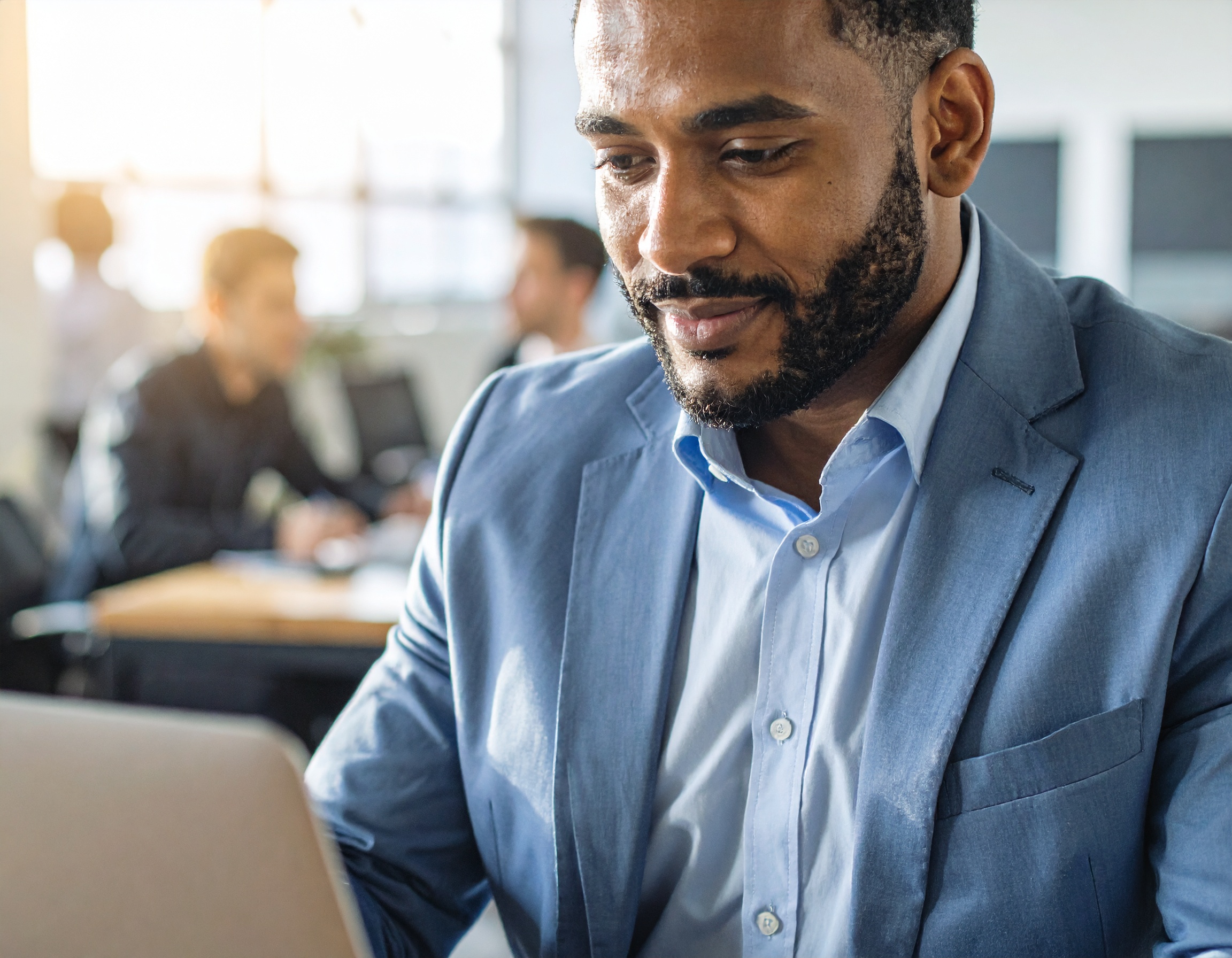 Homem de terno azul claro trabalha concentrado em laptop, em escritório iluminado por luz natural. A sala possui um ambiente colaborativo, com pessoas ao fundo, decoração moderna e cores suaves. A imagem tem foco no rosto do homem, enfatizando expressão de concentração.