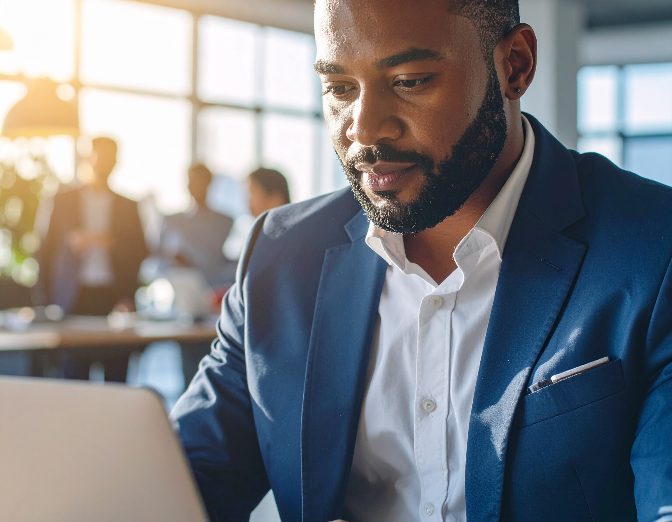 Homem em traje formal azul trabalha em laptop em ambiente de escritório iluminado pelo sol. Composição destaca expressão focada e ambiente profissional moderno com janelas amplas e luz natural suave.