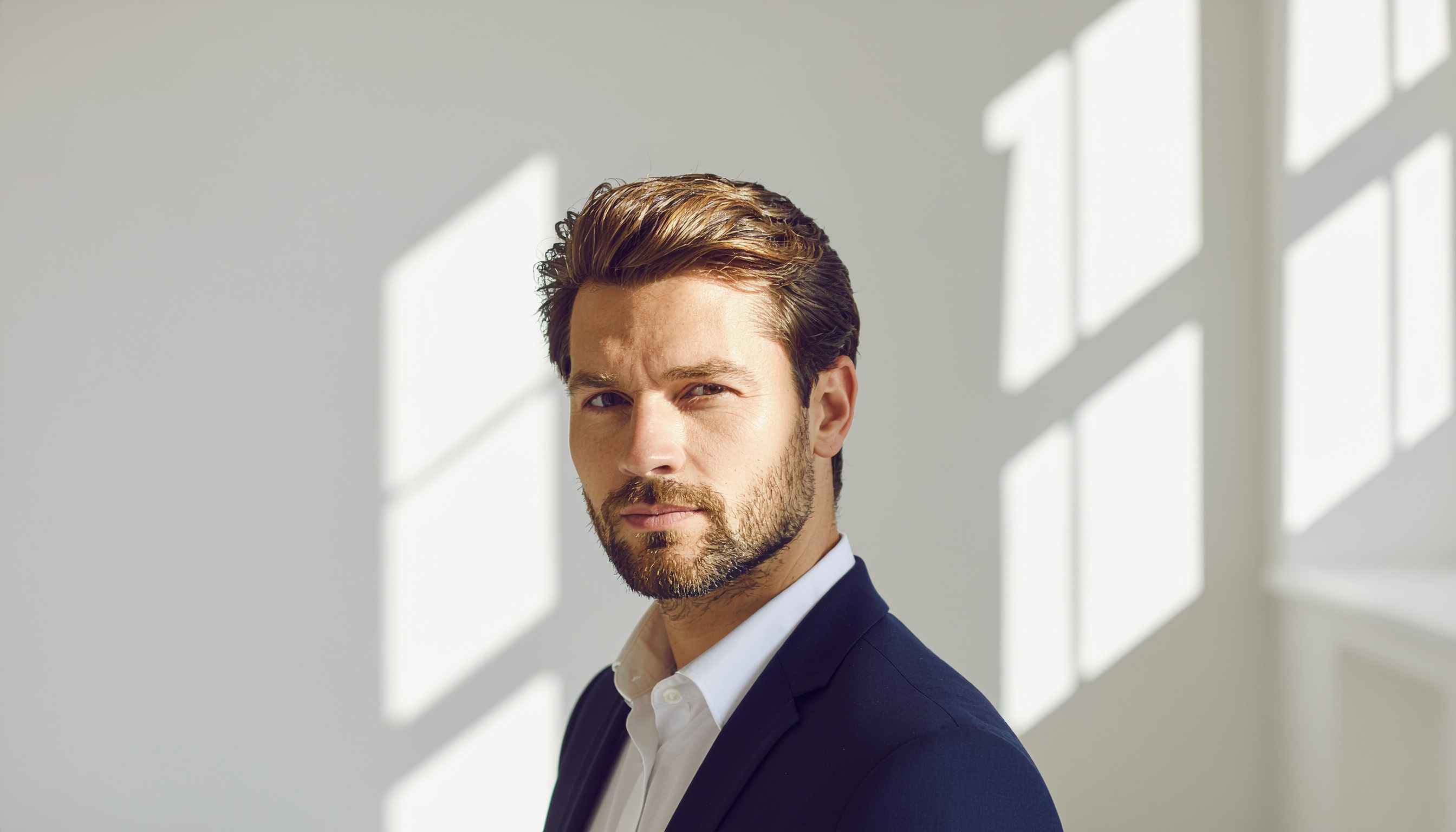 Homem de negócios elegante em terno azul posa em ambiente iluminado por luz natural suave, transmitindo confiança e profissionalismo moderno.