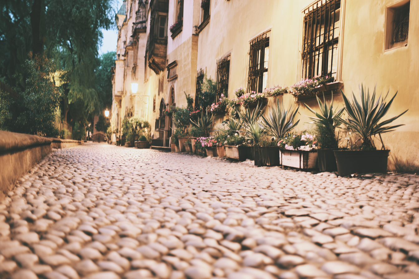 Rua de paralelepípedos à noite com iluminação suave de postes, ladeada por prédios históricos e plantas em vasos, criando uma atmosfera romântica e acolhedora. A perspectiva em ângulo baixo destaca a textura do pavimento, enquanto a luz amarelada reflete nas fachadas antigas. O cenário transmite tranquilidade e charme urbano, com foco suave que adiciona um toque de mistério.