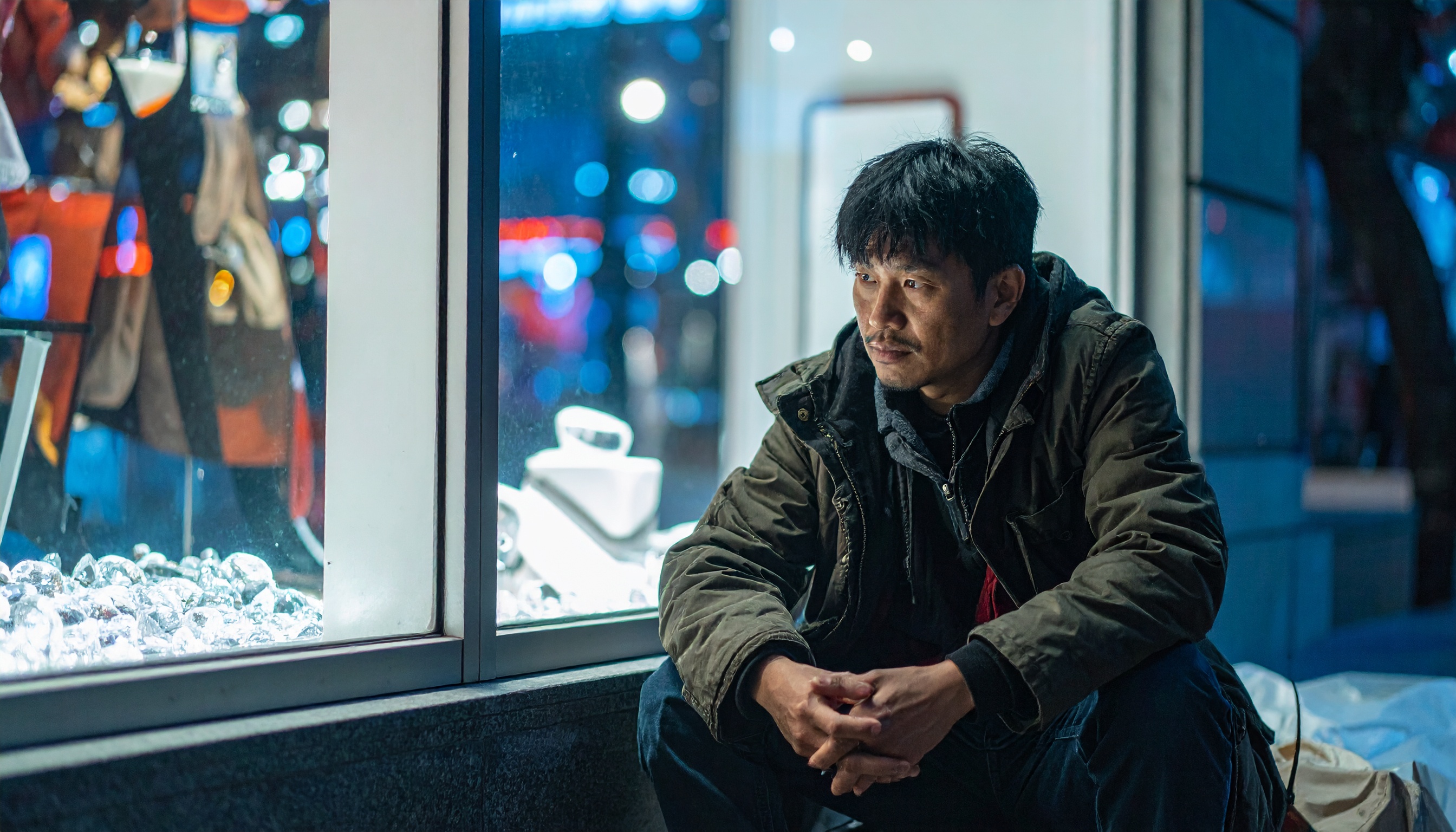 A contemplative man sits beside a brightly lit shop window at night