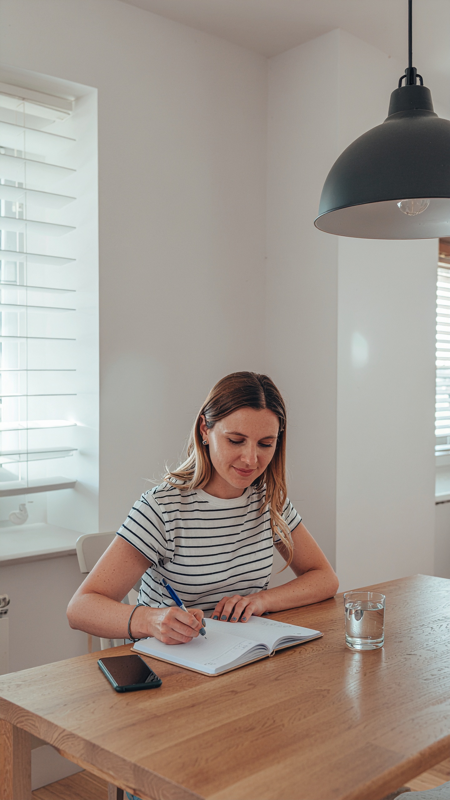 A woman in a striped shirt writes in a notebook at a wooden table