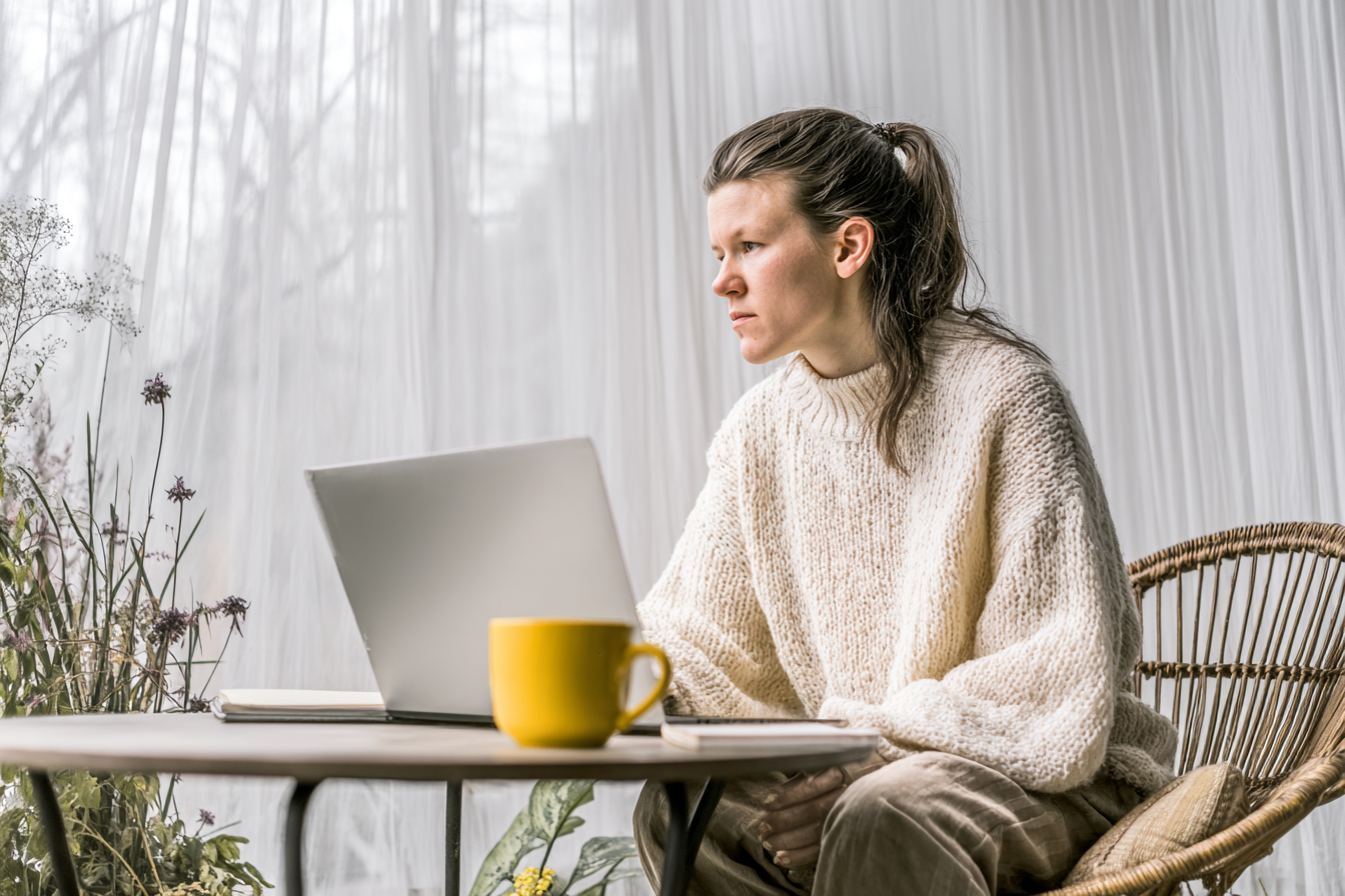 Mulher concentrada trabalha remotamente em laptop, sentada em uma cadeira de vime em sala iluminada por luz natural suave, com xícara de café amarela e plantas ao fundo, transmitindo tranquilidade e foco.