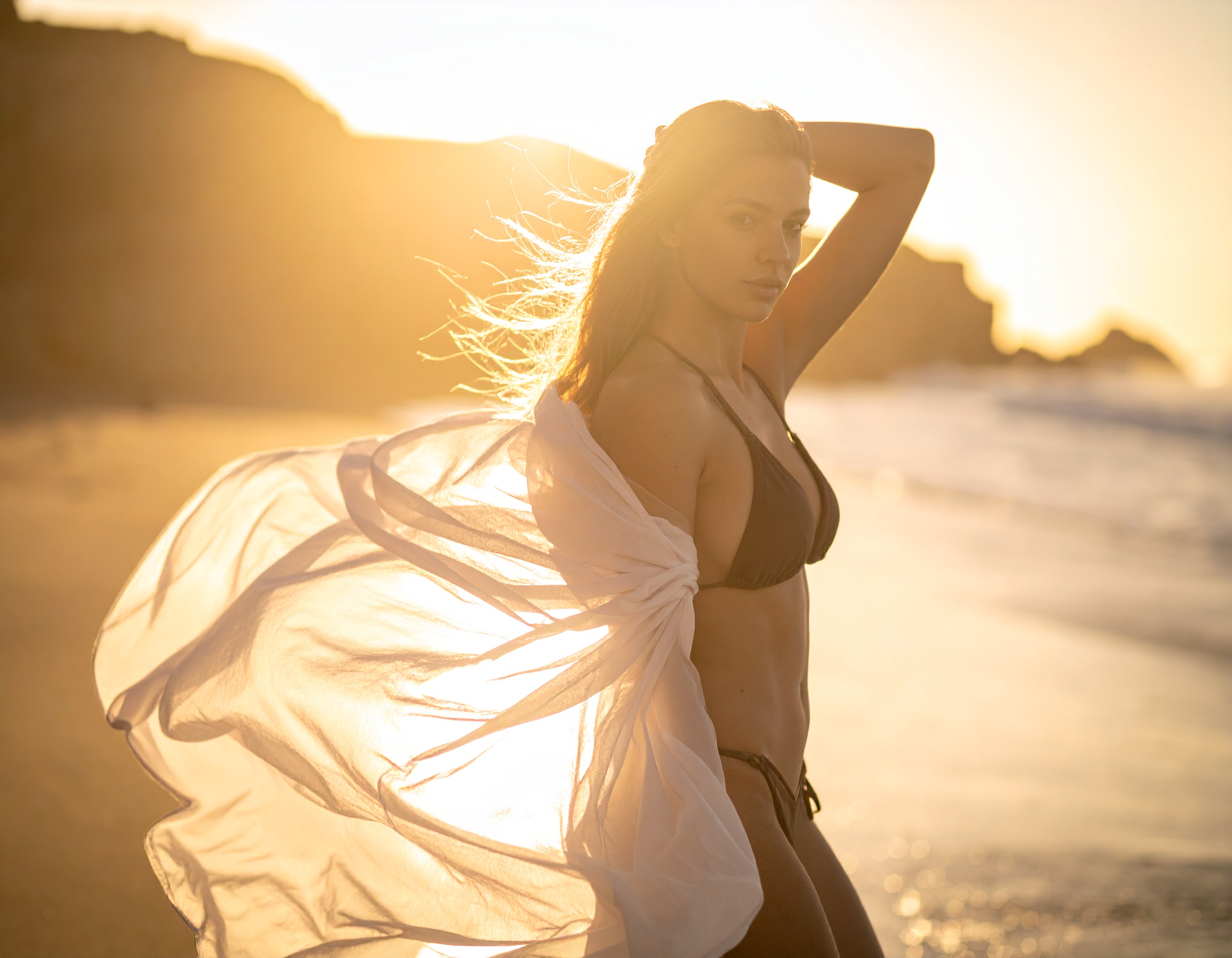 A woman in a black bikini stands on a sunlit beach, wrapped in a flowing white fabric