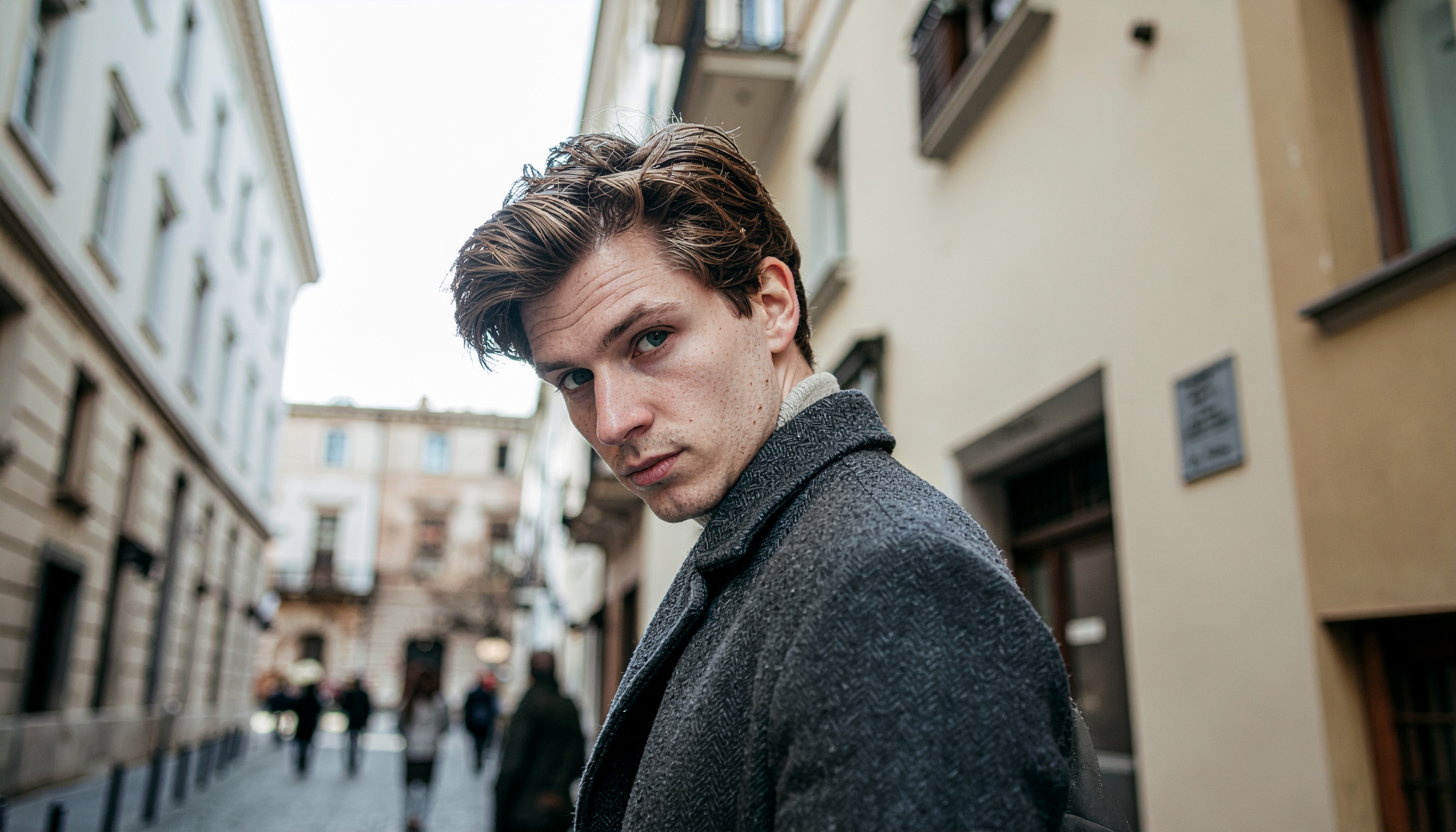 A man in a grey coat stands confidently in a narrow urban street