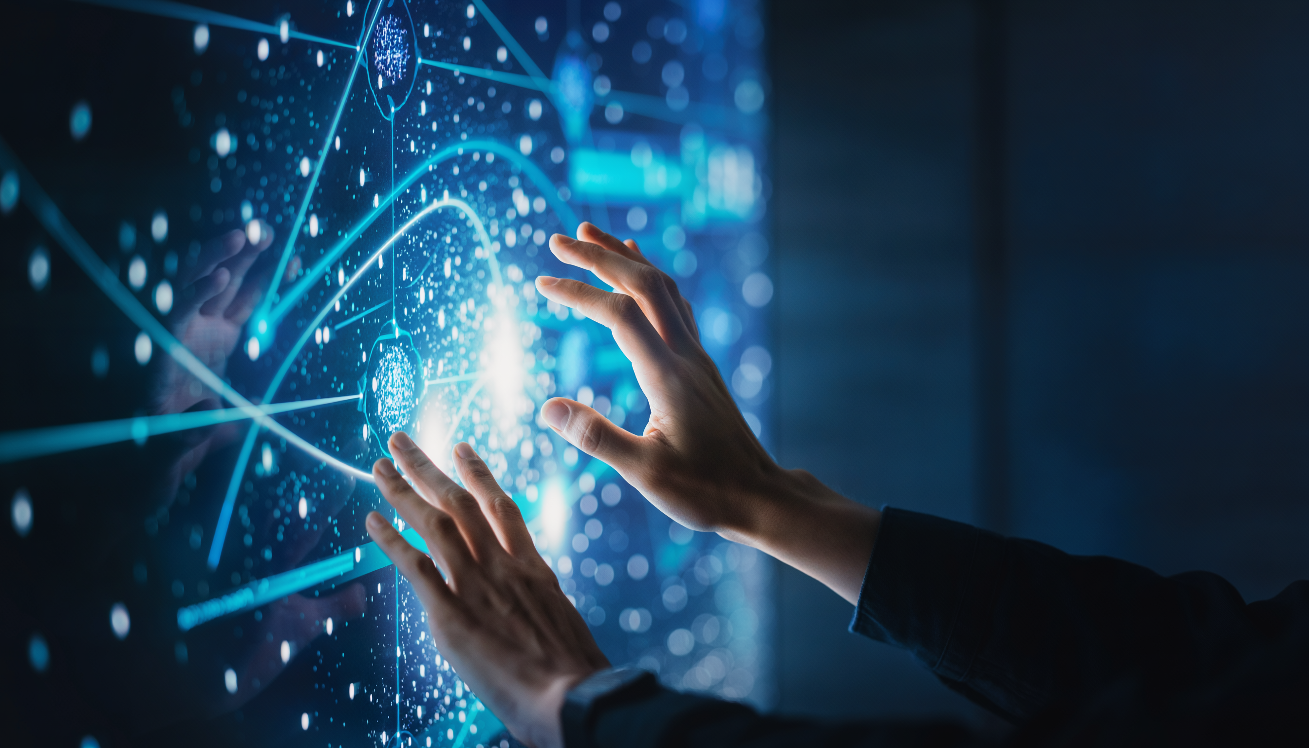 Mãos interagindo com tela digital futurista iluminada em tons de azul, exibindo gráficos complexos e linhas de conectividade, criando uma sensação de tecnologia avançada e imersão em um ambiente de alta tecnologia. A composição enfatiza a interação humana com interfaces digitais por meio de um foco suave nas mãos e nos elementos gráficos, sugerindo inovação e conectividade global.