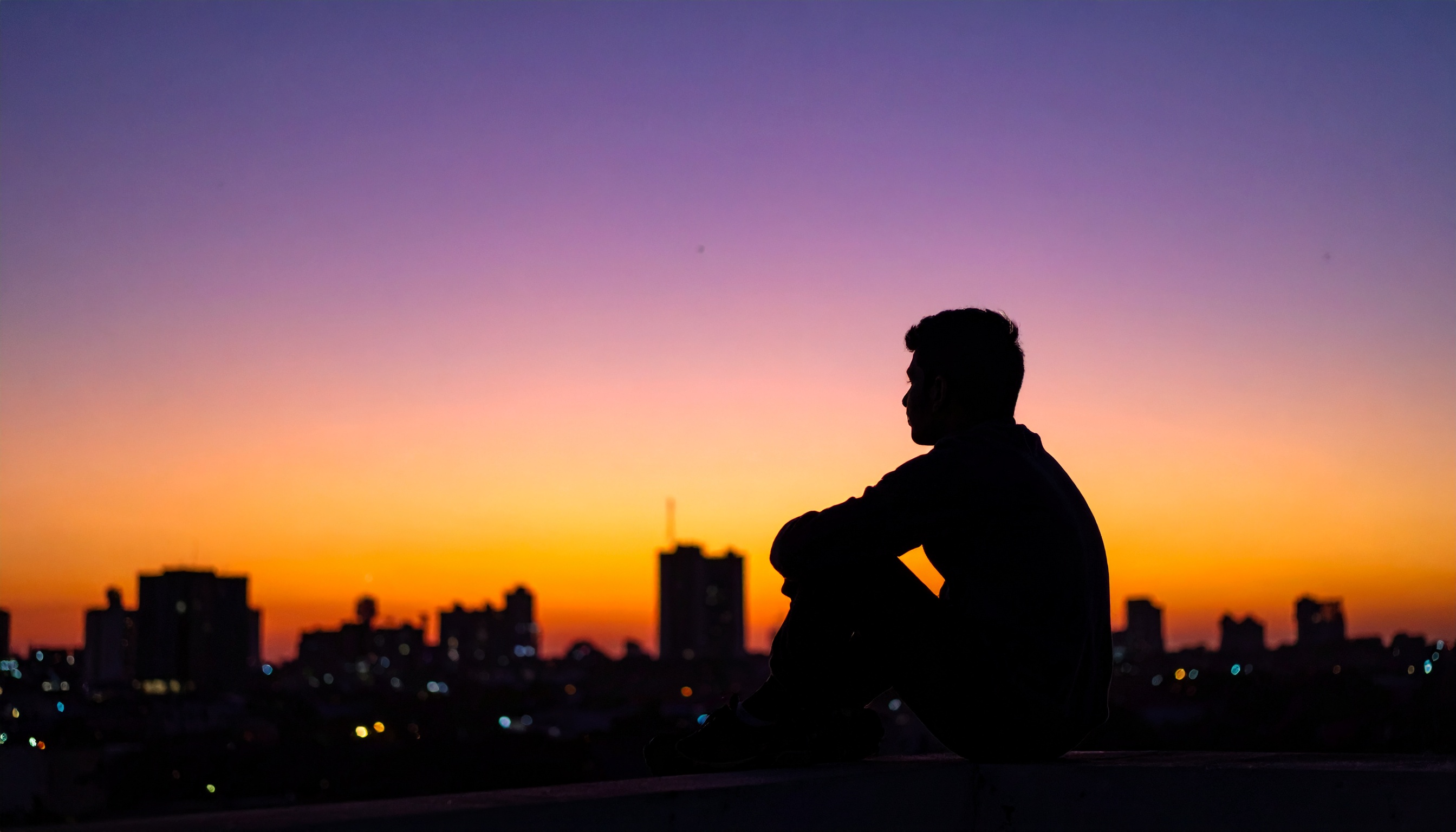A silhouette of a person sits against a vibrant sunset over a city skyline