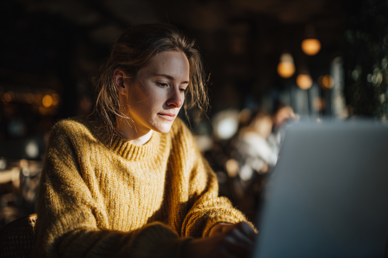 Mulher jovem em suéter amarelo digitando em laptop em ambiente de café, com iluminação suave destacando seu rosto e fundo desfocado, criando uma atmosfera acolhedora e de concentração.
