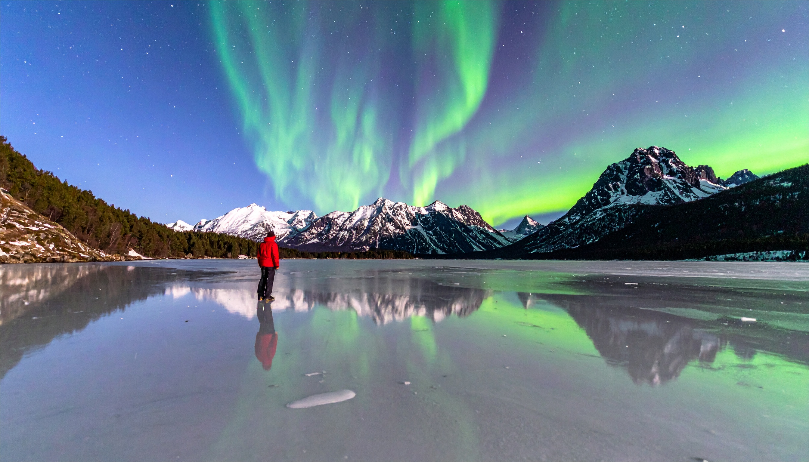 A person in a red jacket stands on a frozen lake under vibrant auroras