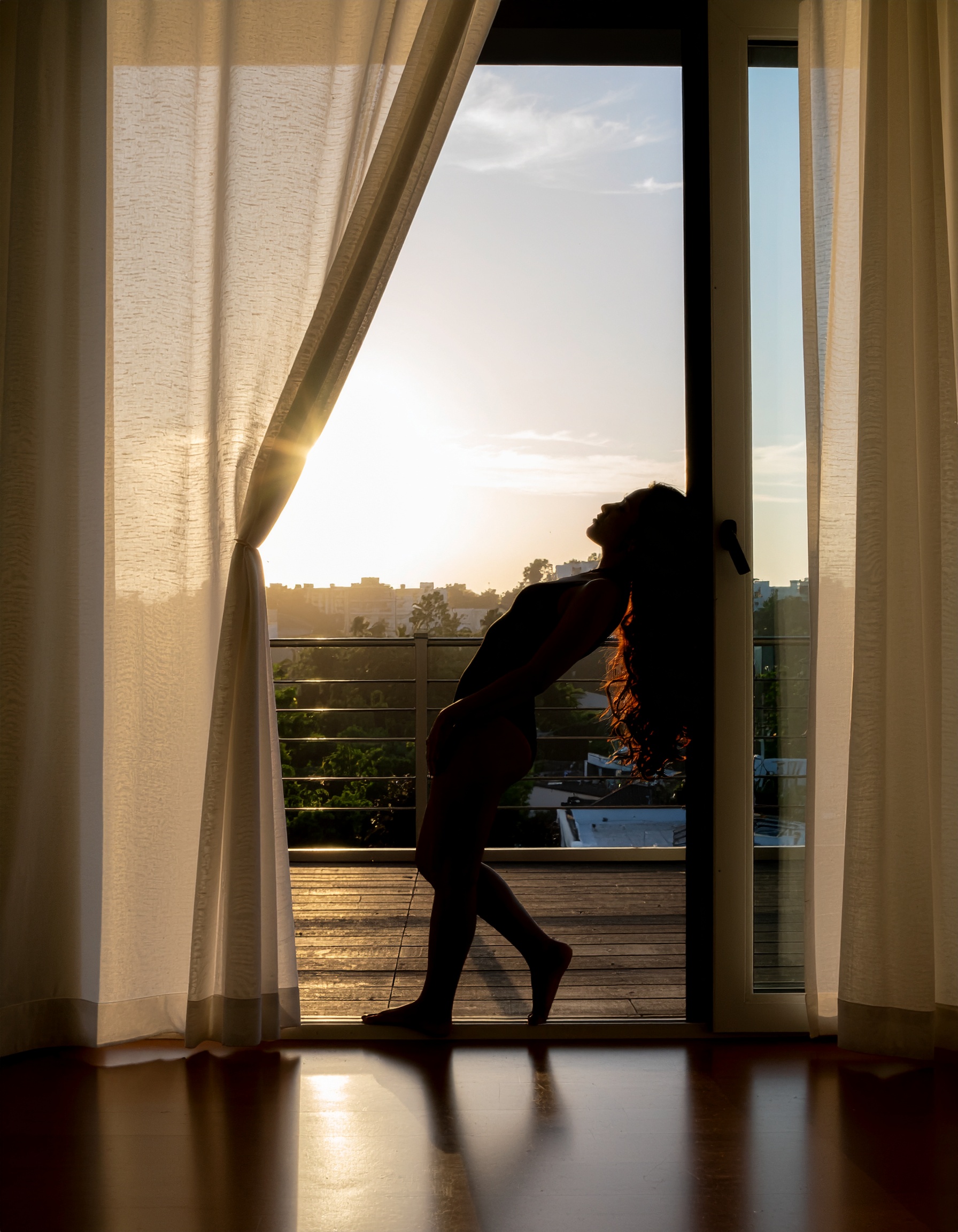 A silhouette of a person stands gracefully against a backdrop of sheer curtains and a bright sunset