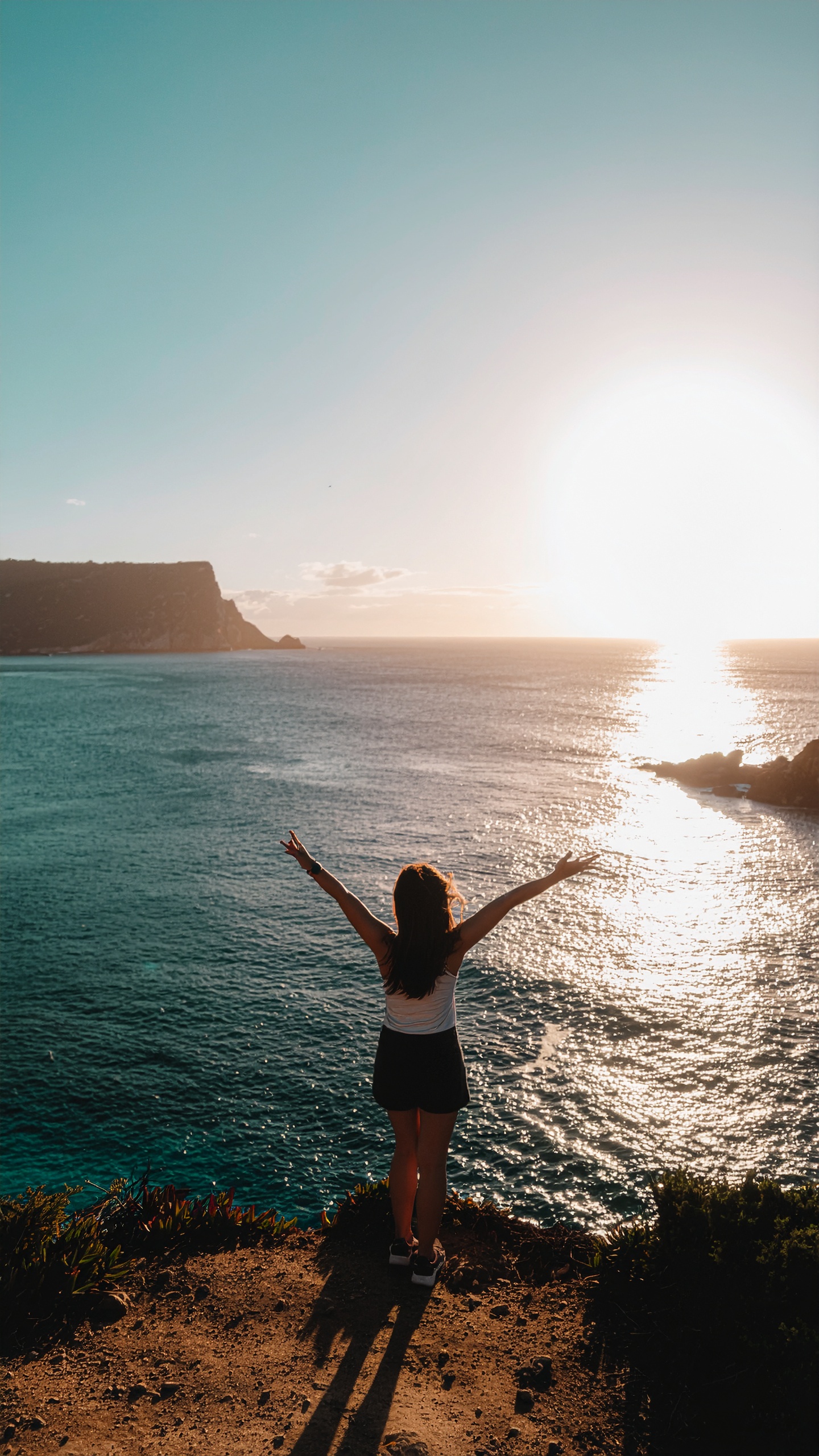 Uma mulher de braços abertos contempla o pôr do sol sobre um oceano cintilante, no topo de um penhasco, transmitindo liberdade e serenidade.