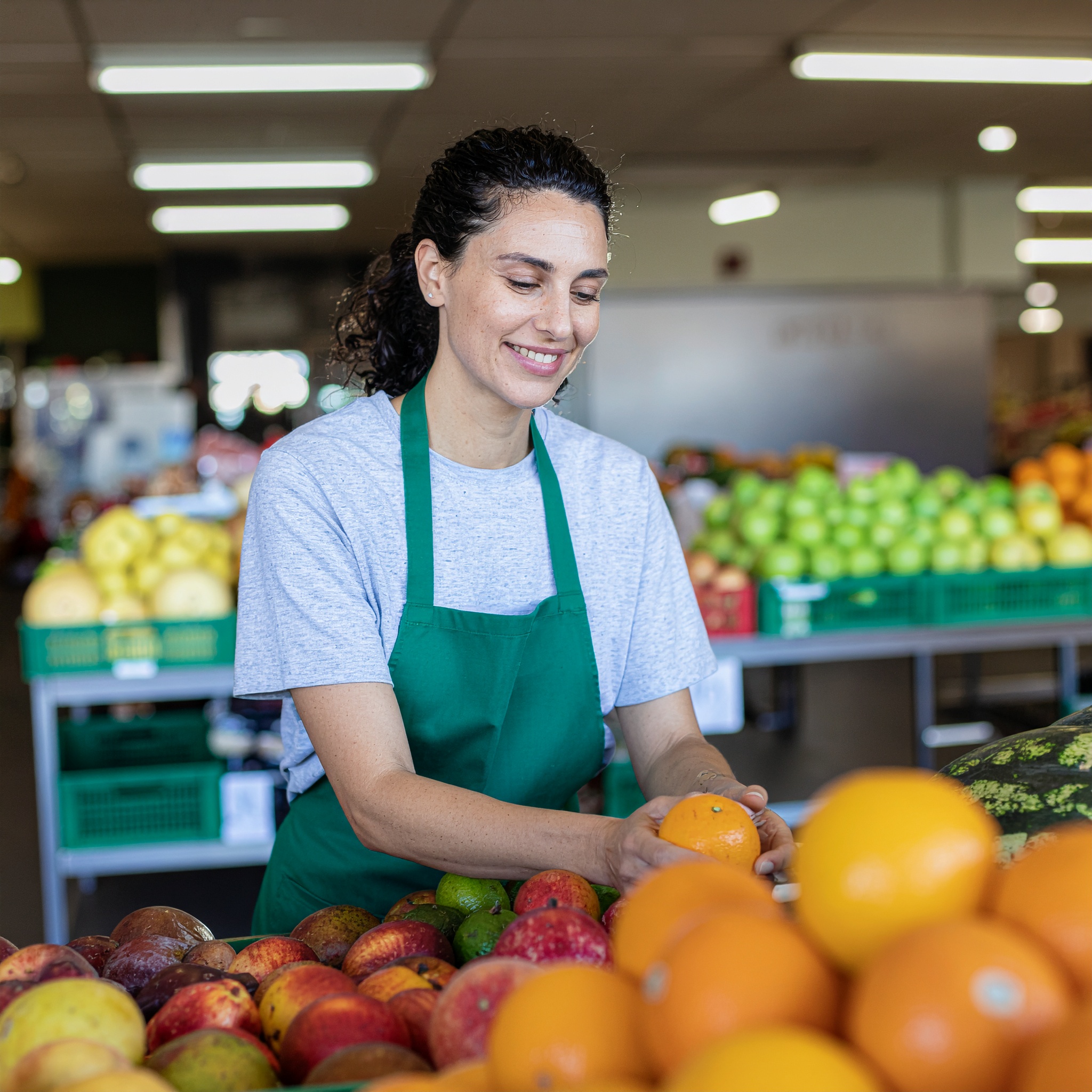 Mulher sorridente com avental verde organizando frutas frescas em um mercado bem iluminado. A composição destaca maçãs, laranjas e melancias em primeiro plano, criando uma atmosfera acolhedora e vibrante. A iluminação suave realça a variedade de cores das frutas sob um ângulo direto que mantém o foco nítido na cena.