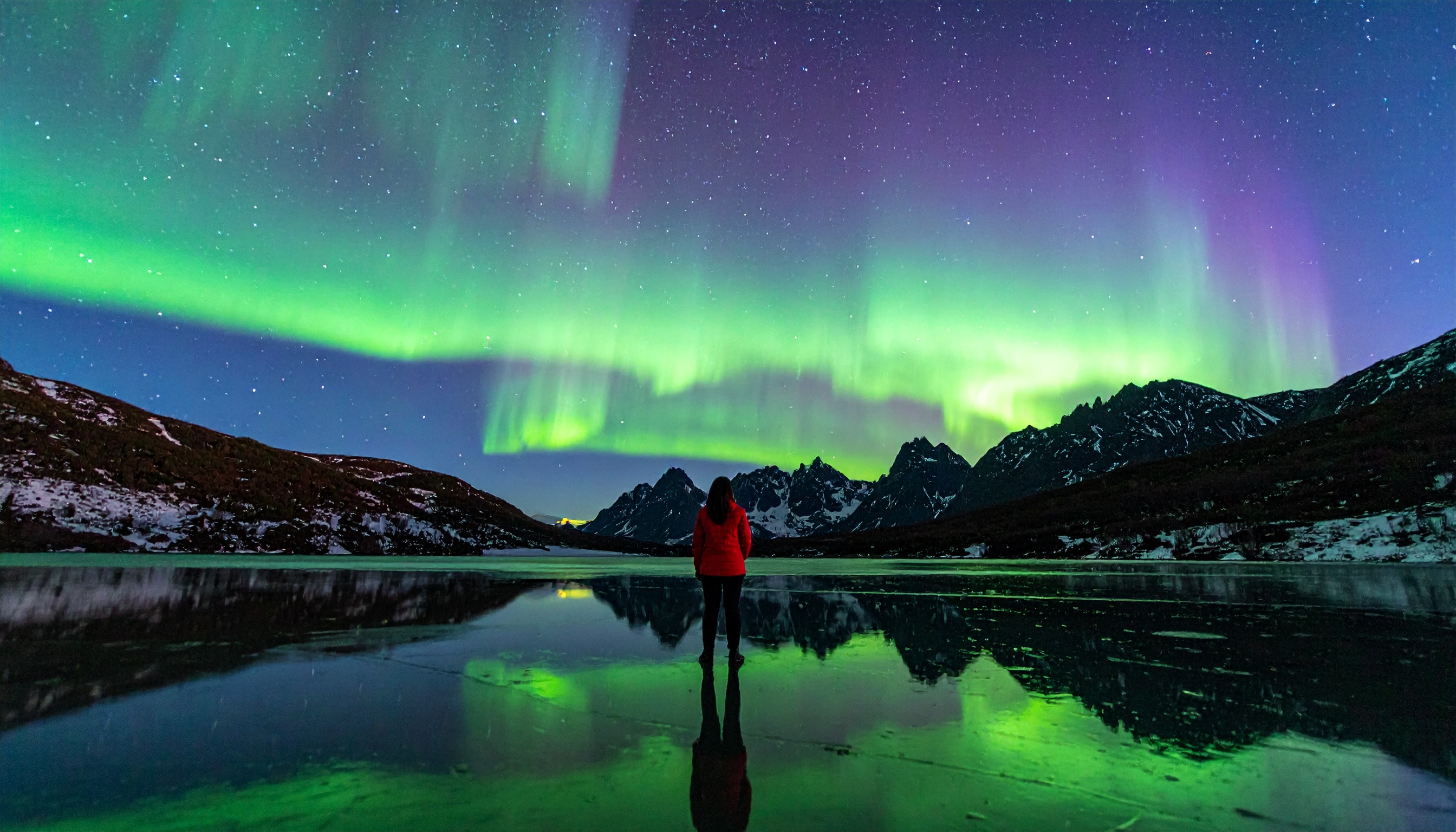 A person in a red jacket stands by a serene icy lake under vibrant Northern Lights