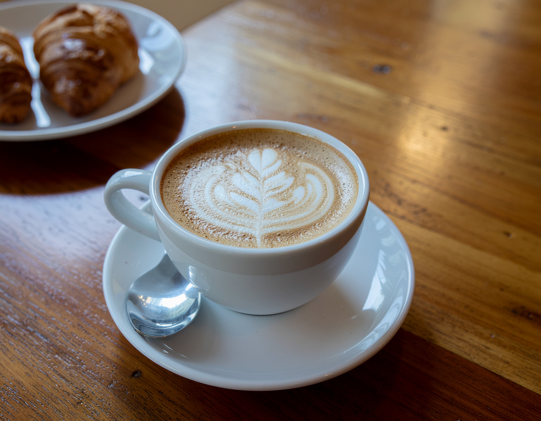 Xícara de café com latte art em uma mesa de madeira, ao lado de croissants frescos. Ideal para momentos de relaxamento ou encontros em cafés aconchegantes. A espuma foi esculpida em um desenho delicado, destacando a habilidade barista. Perfeito para acompanhar um café da manhã ou lanche da tarde. Oferece uma experiência visual e gustativa atraente, valorizando encontros sociais.