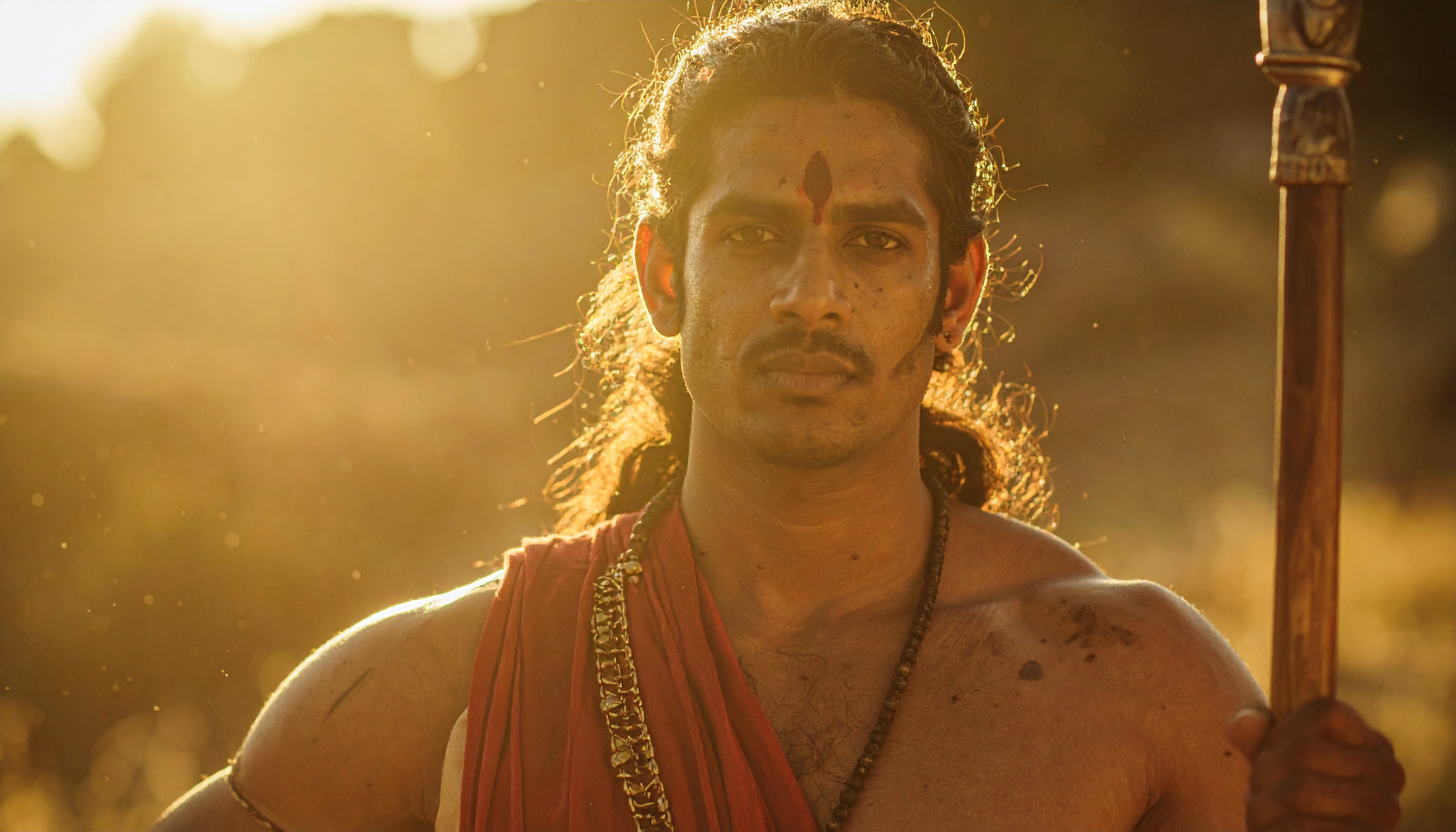 A man dressed in traditional attire stands against a golden sunset backdrop