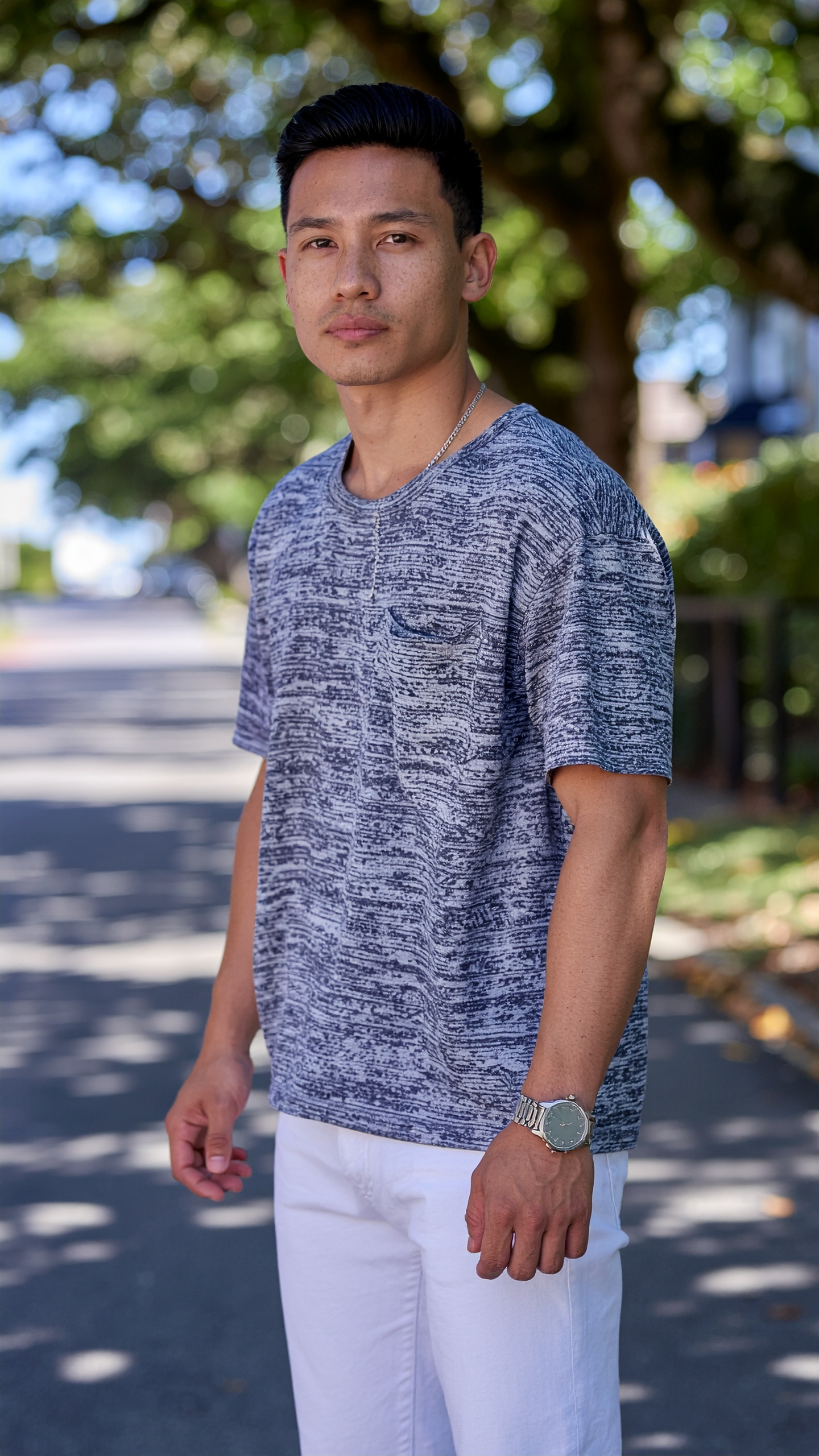 A man stands confidently on a sunlit street wearing a casual grey patterned t-shirt and white pants