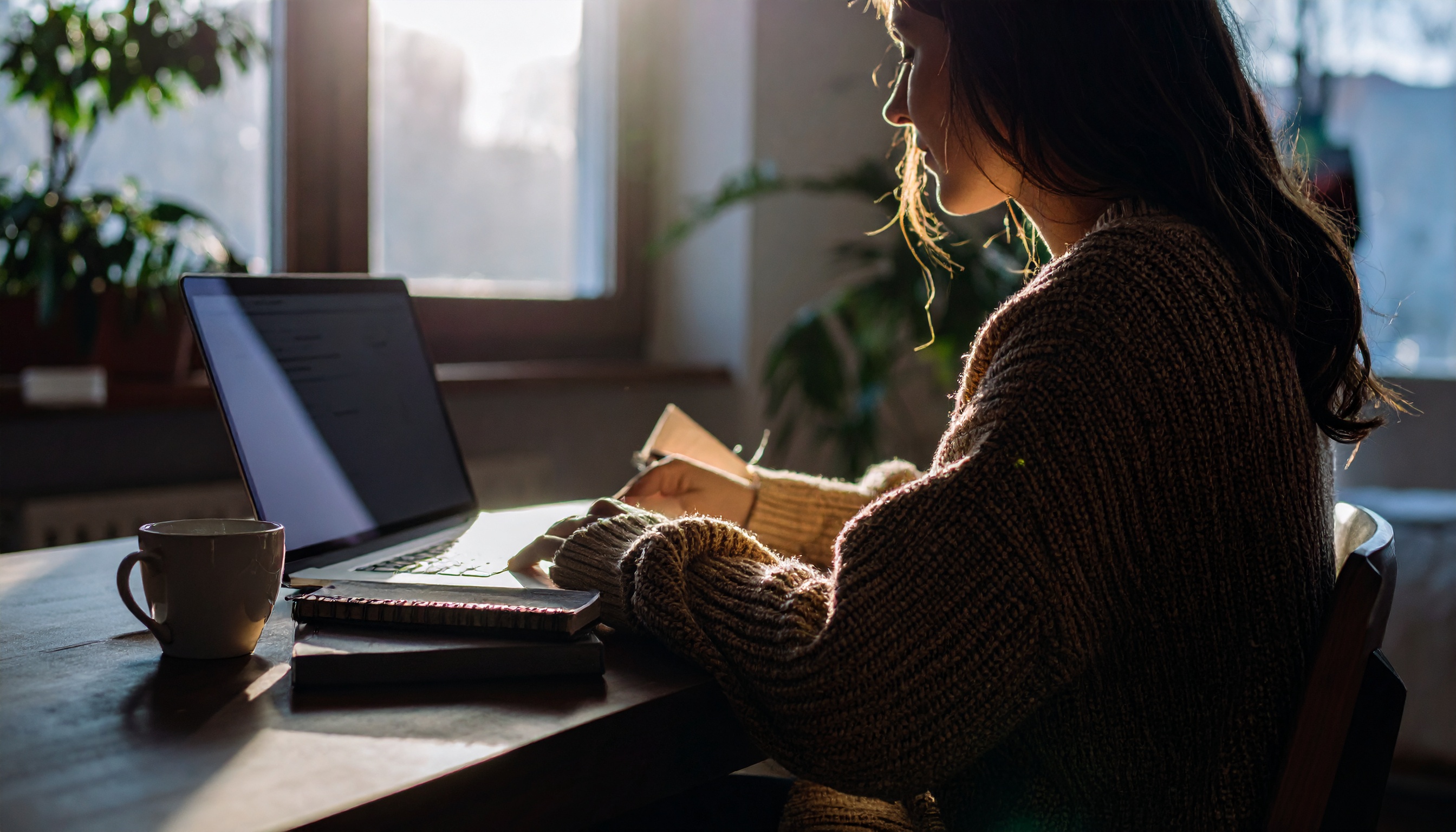 Mulher escrevendo em caderno ao lado de laptop em mesa de madeira, iluminada pela luz suave da manhã, transmitindo foco e concentração em ambiente aconchegante.