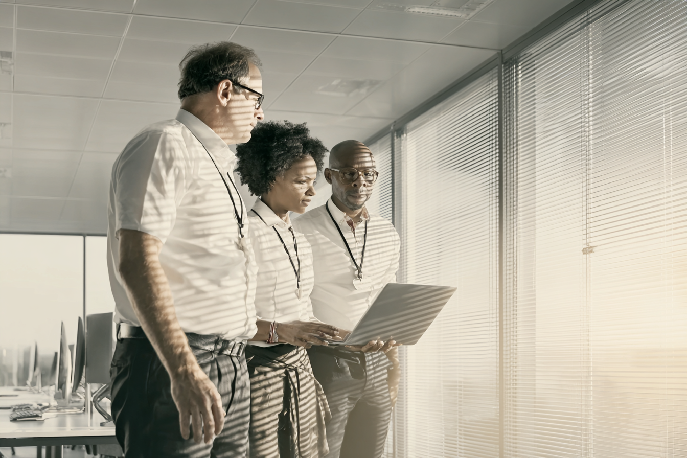 Três profissionais em um ambiente de escritório moderno observam atentamente um laptop. A luz suave do sol atravessa as persianas, criando padrões de sombra nas paredes e nos indivíduos. Eles estão vestidos formalmente, em pé próximos a uma janela ampla, que oferece uma sensação de abertura e colaboração. A composição é equilibrada, com foco nítido nas expressões concentradas dos participantes, sugerindo uma reunião ou análise de dados importante.