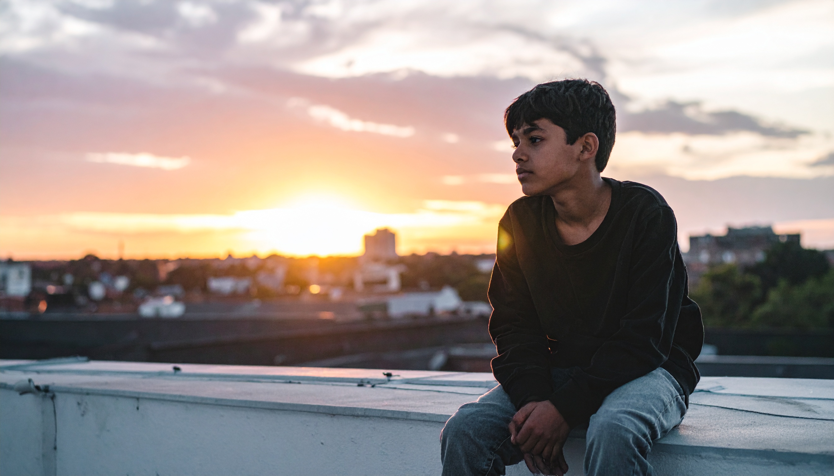 A young person sits on a rooftop at sunset, gazing thoughtfully into the distance