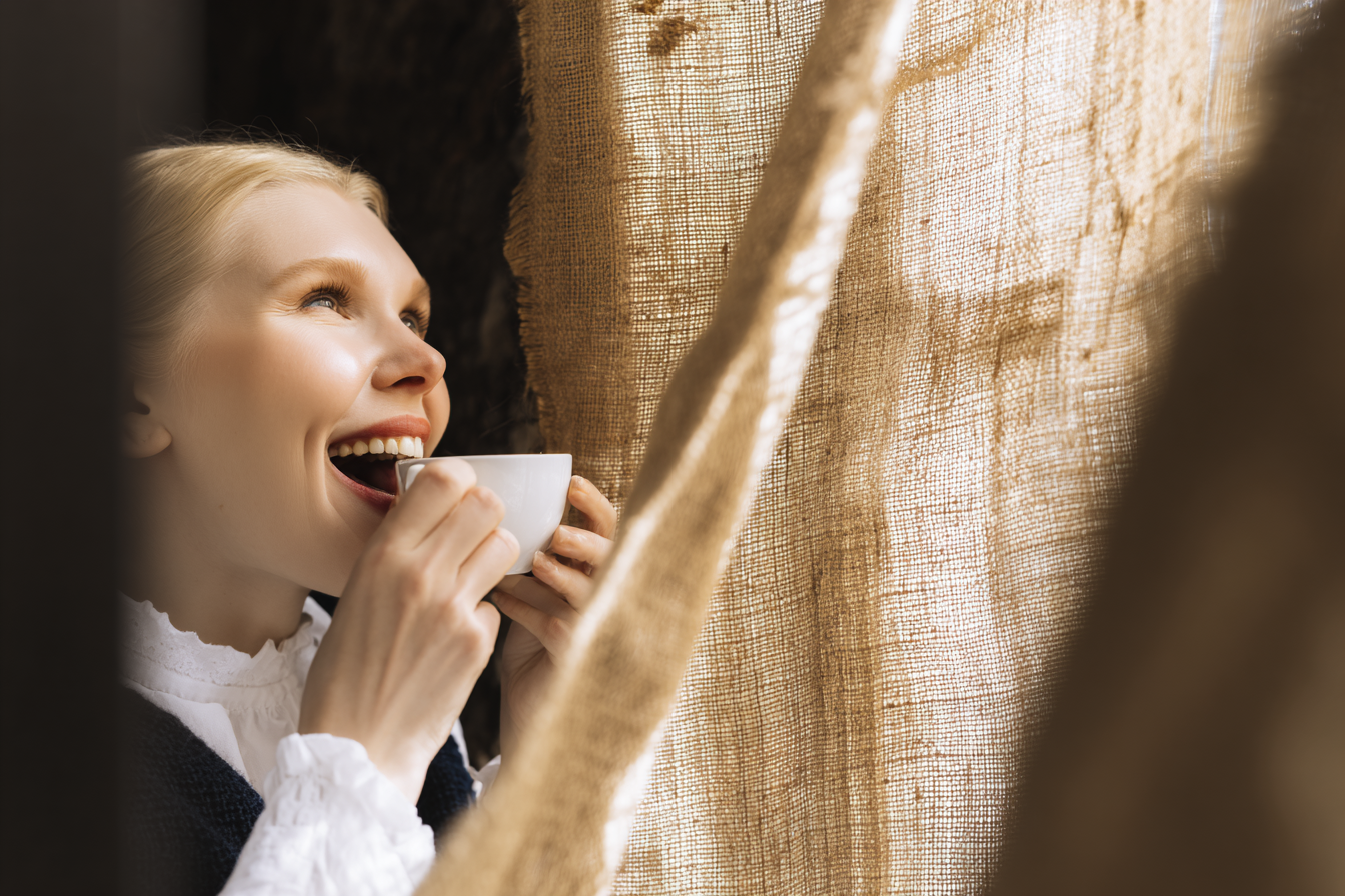 Mulher sorridente segurando uma xícara de café perto de uma cortina de linho, com iluminação suave destacando seu rosto. O ambiente tem uma atmosfera acolhedora e serena, focado no prazer de um momento tranquilo. A composição destaca a textura da cortina e a expressão de felicidade da mulher, com um ângulo lateral que captura a luz natural iluminando suavemente a cena.