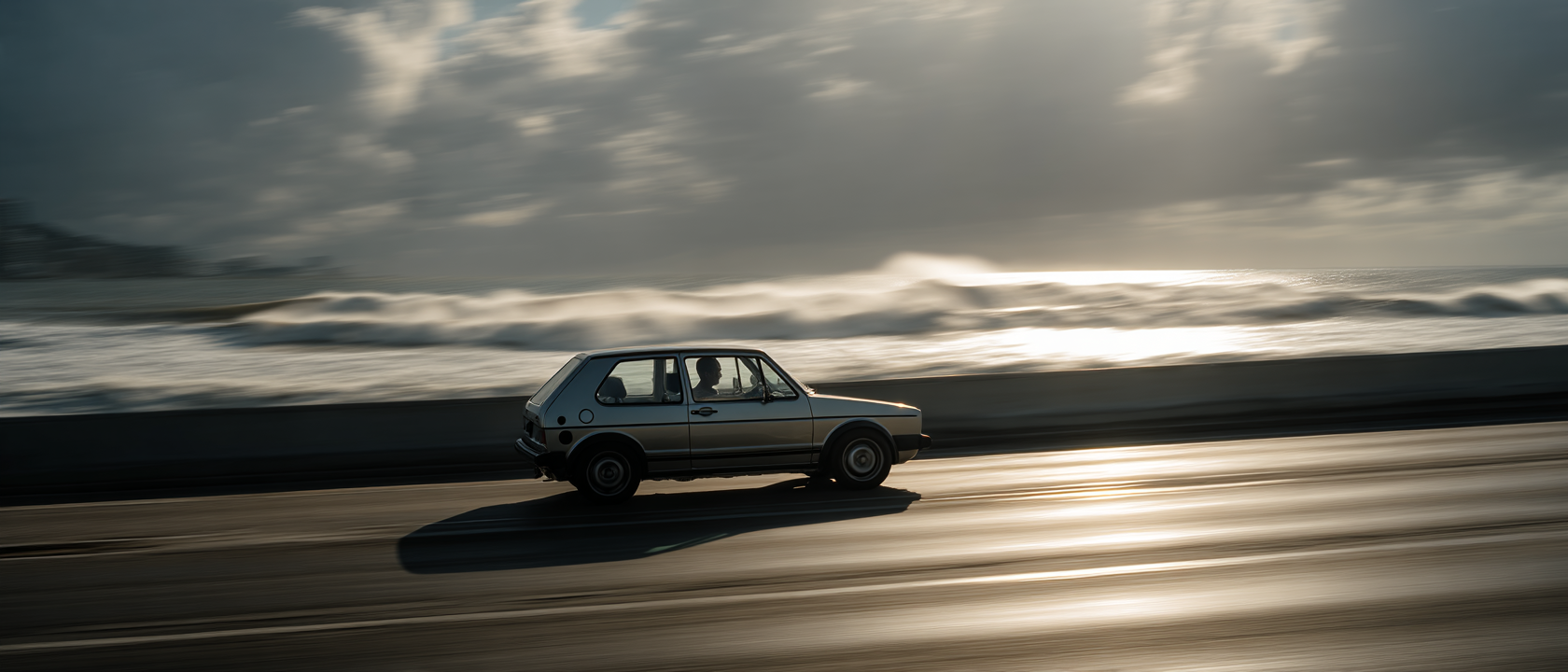 Um carro compacto de cor clara passando rapidamente por uma estrada à beira-mar, com ondas quebrando ao fundo sob um céu nublado. A imagem captura a sensação de movimento e liberdade em um cenário costeiro.