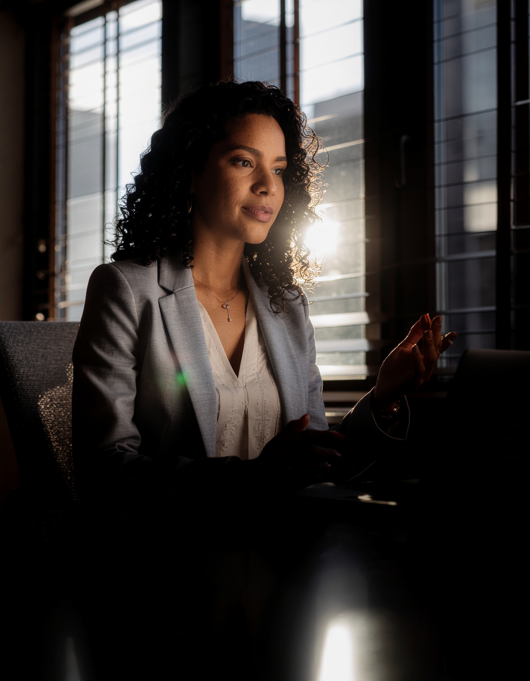 Mulher em ambiente de escritório iluminado por luz natural intensa, refletida através de janelas, criando contraste dramático. Ela está vestindo blazer cinza, com expressão contemplativa, capturada em perfil. A cena transmite profissionalismo e serenidade, enquanto o foco está suavemente centrado nela, com profundidade de campo reduzida acentuando o ambiente elegante.