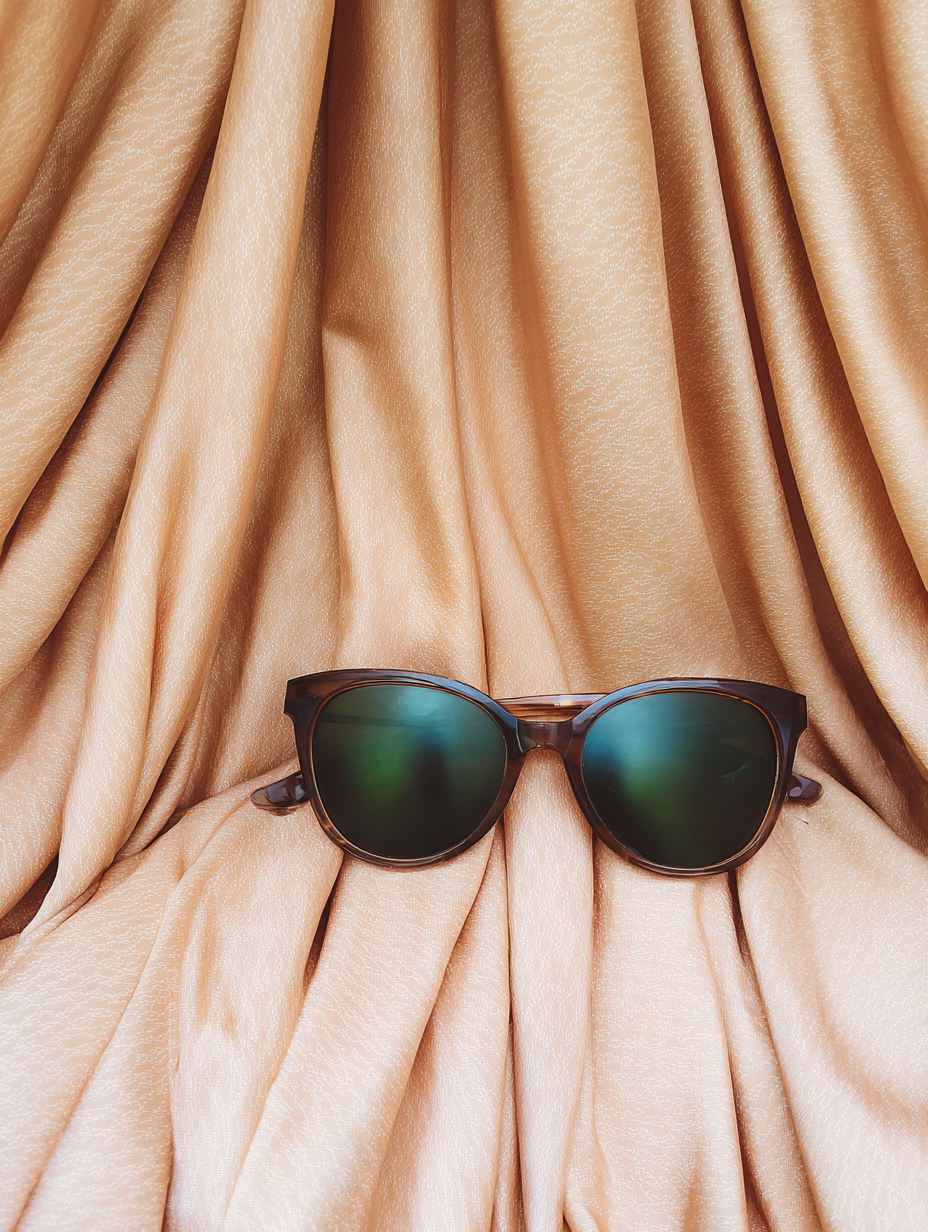 Óculos de sol de armação marrom sobre tecido dourado drapeado, com lentes verdes refletivas. Iluminação suave destaca as nuances do tecido e a textura das lentes, criando uma composição harmônica e elegante.