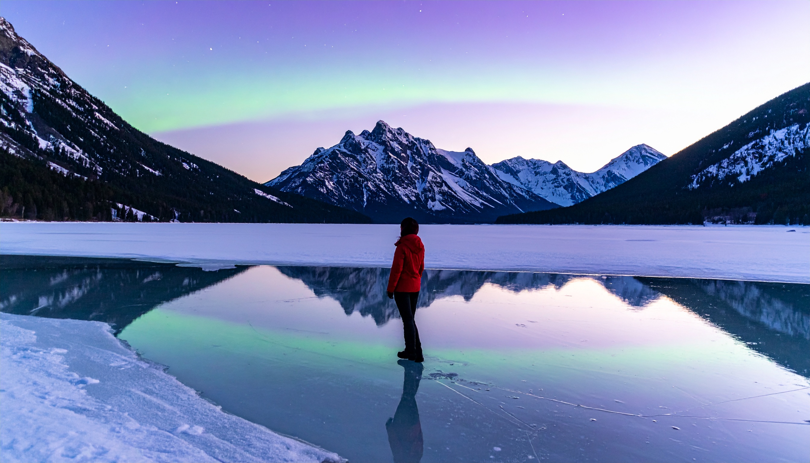 A person in a red jacket stands on a frozen lake with majestic mountains in the background