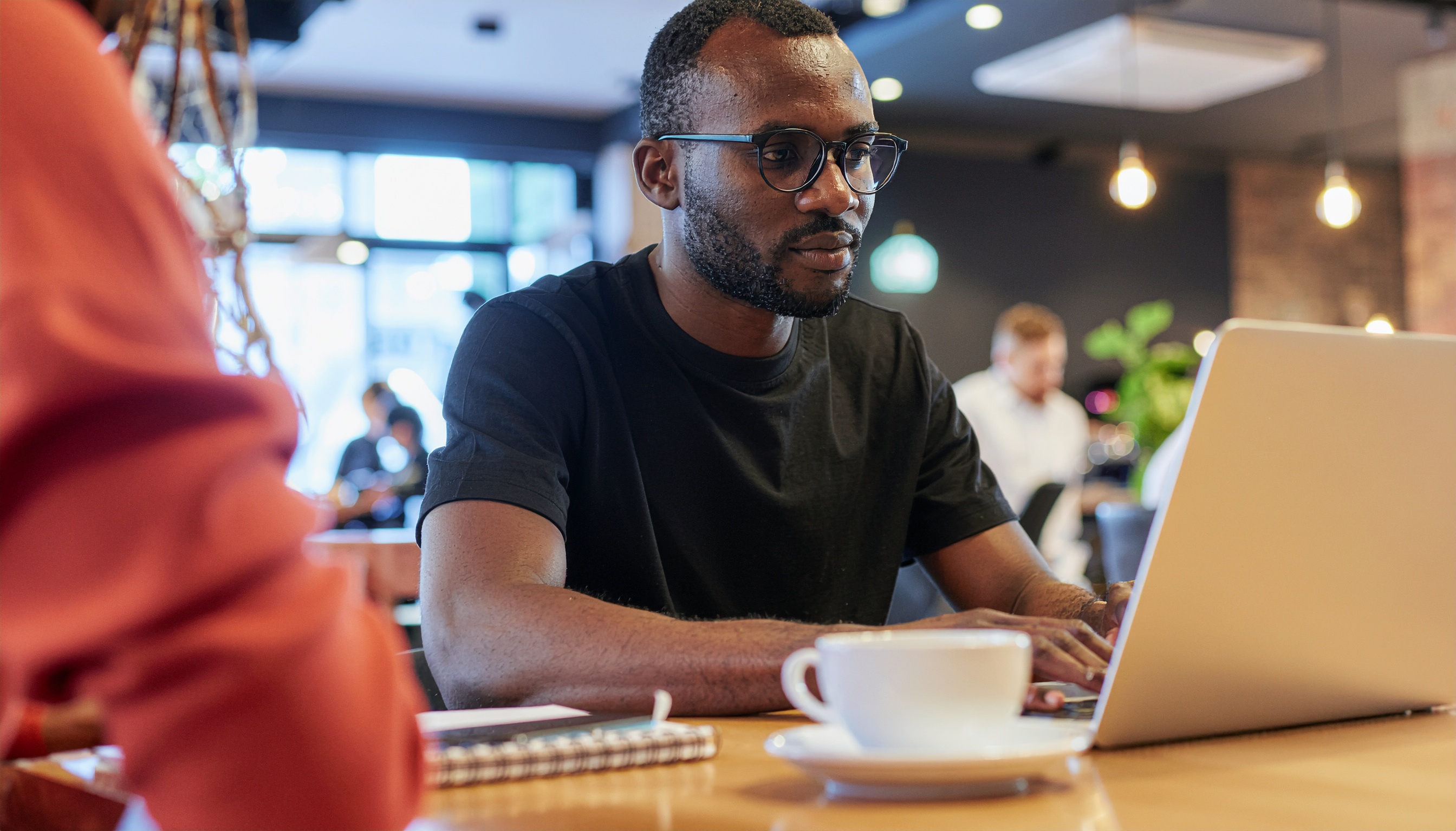 Homem usando óculos trabalhando em laptop em um café moderno, iluminação suave e ambiente descontraído. Cena focada, com fundo desfocado mostrando pessoas ocupadas.
