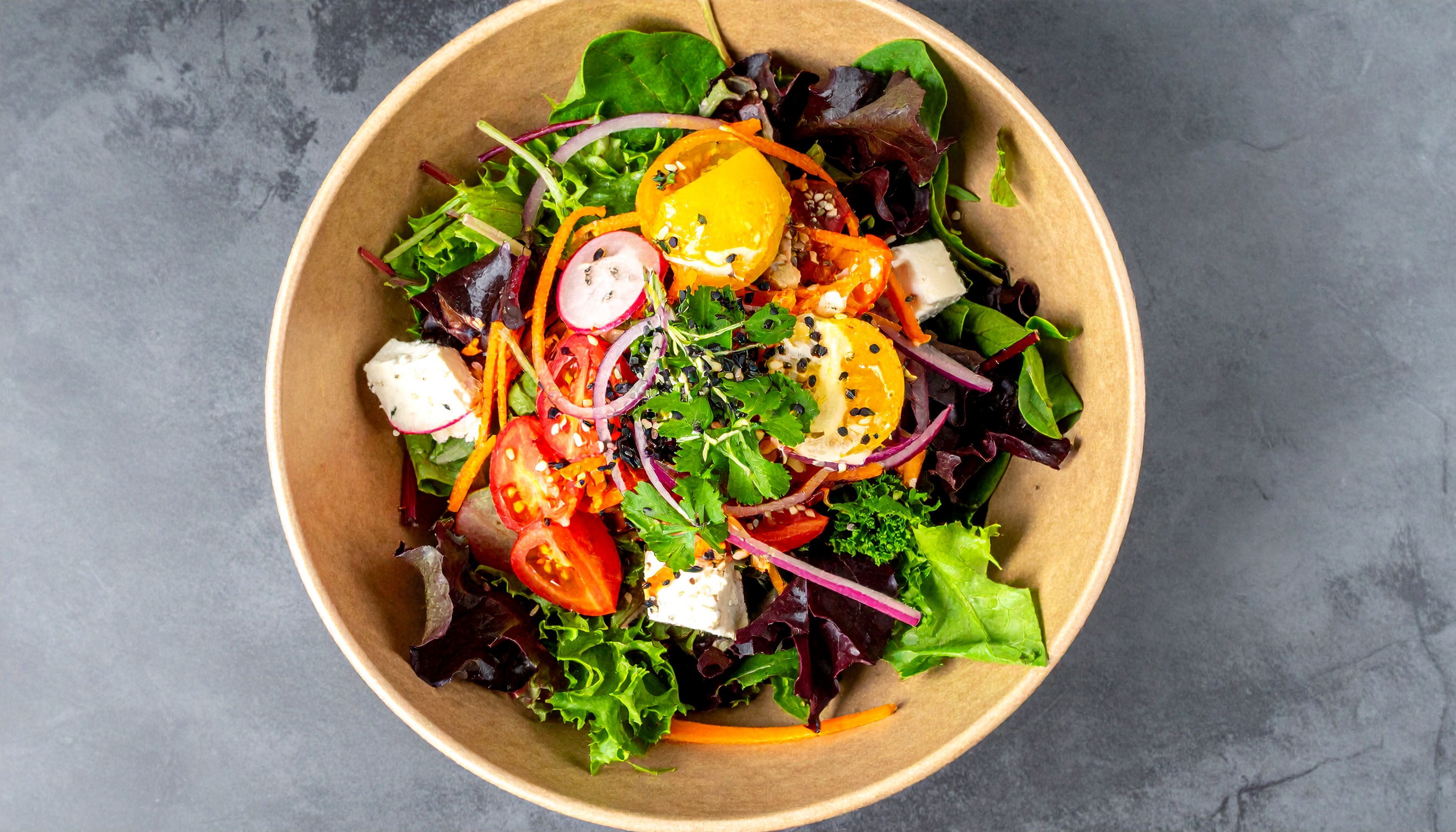 Salada vibrante em tigela marrom com folhas verdes, tomates, cebolas roxas, cenouras e queijo, destacando cores vivas e texturas frescas sobre fundo cinza.
