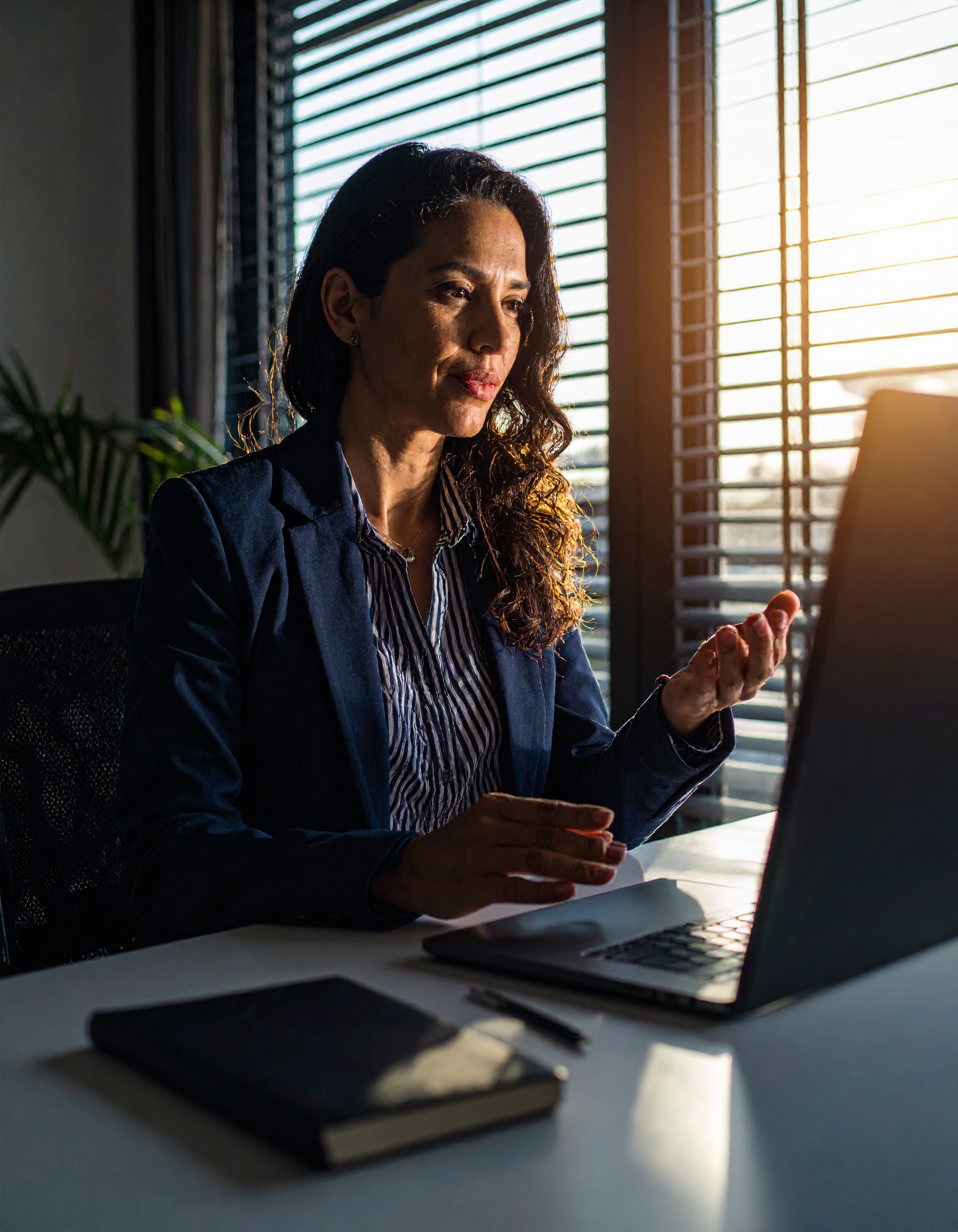 Mulher em traje formal está sentada em escritório iluminado pelo sol poente, usando laptop sobre mesa branca com caderno ao lado. A luz dourada atravessa persianas, criando um contraste suave e uma atmosfera profissional. O enquadramento é frontal com foco nítido na expressão concentrada da mulher.