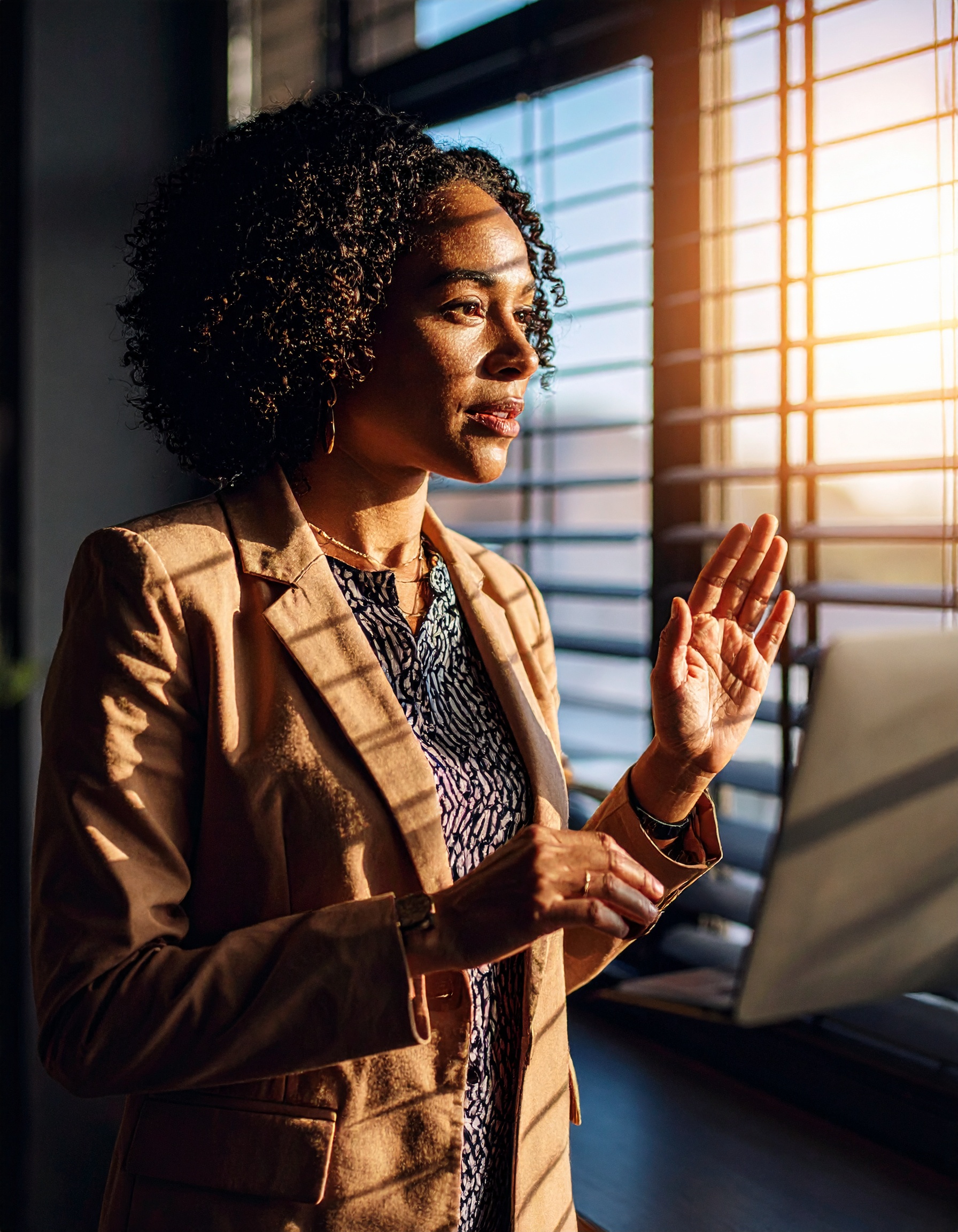 Mulher em traje formal olha pela janela com persianas, iluminada pelo sol poente, criando sombras suaves no ambiente. O cenário sugere um escritório, com luz dourada adicionando um tom caloroso e contemplativo à cena. A composição destaca o perfil da mulher, com foco preciso, capturando uma expressão de reflexão e introspecção.