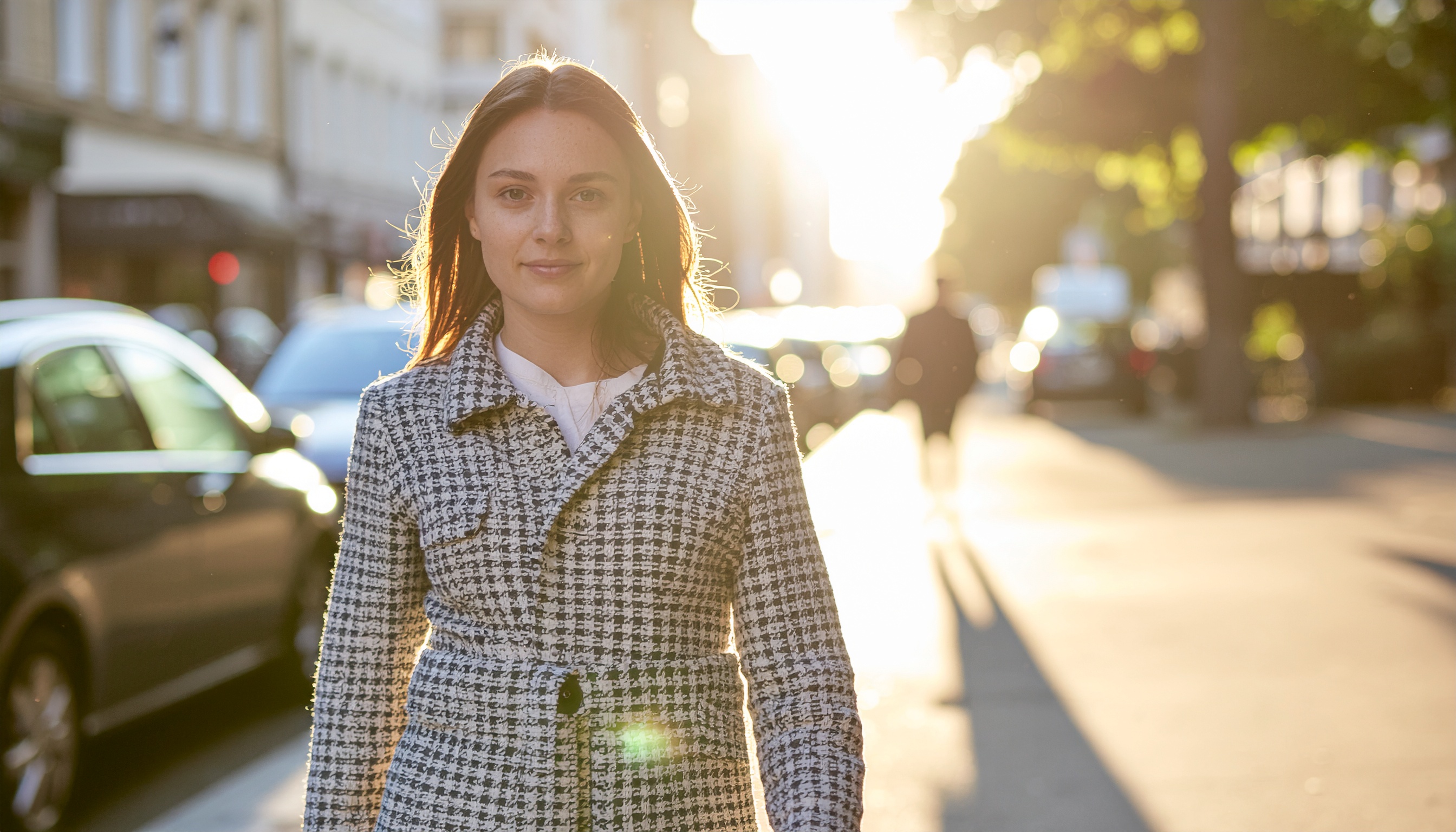Mulher caminhando em uma rua urbana ensolarada com um casaco xadrez elegante. O casaco de padrão clássico é ideal para ocasiões formais e uso cotidiano, oferecendo estilo e conforto. A cena destaca a luz dourada do pôr do sol, criando um ambiente acolhedor e sofisticado. Perfeito para passeios ao entardecer ou encontros casuais em locais urbanos. A combinação de moda e cenário urbano transmite modernidade e elegância.