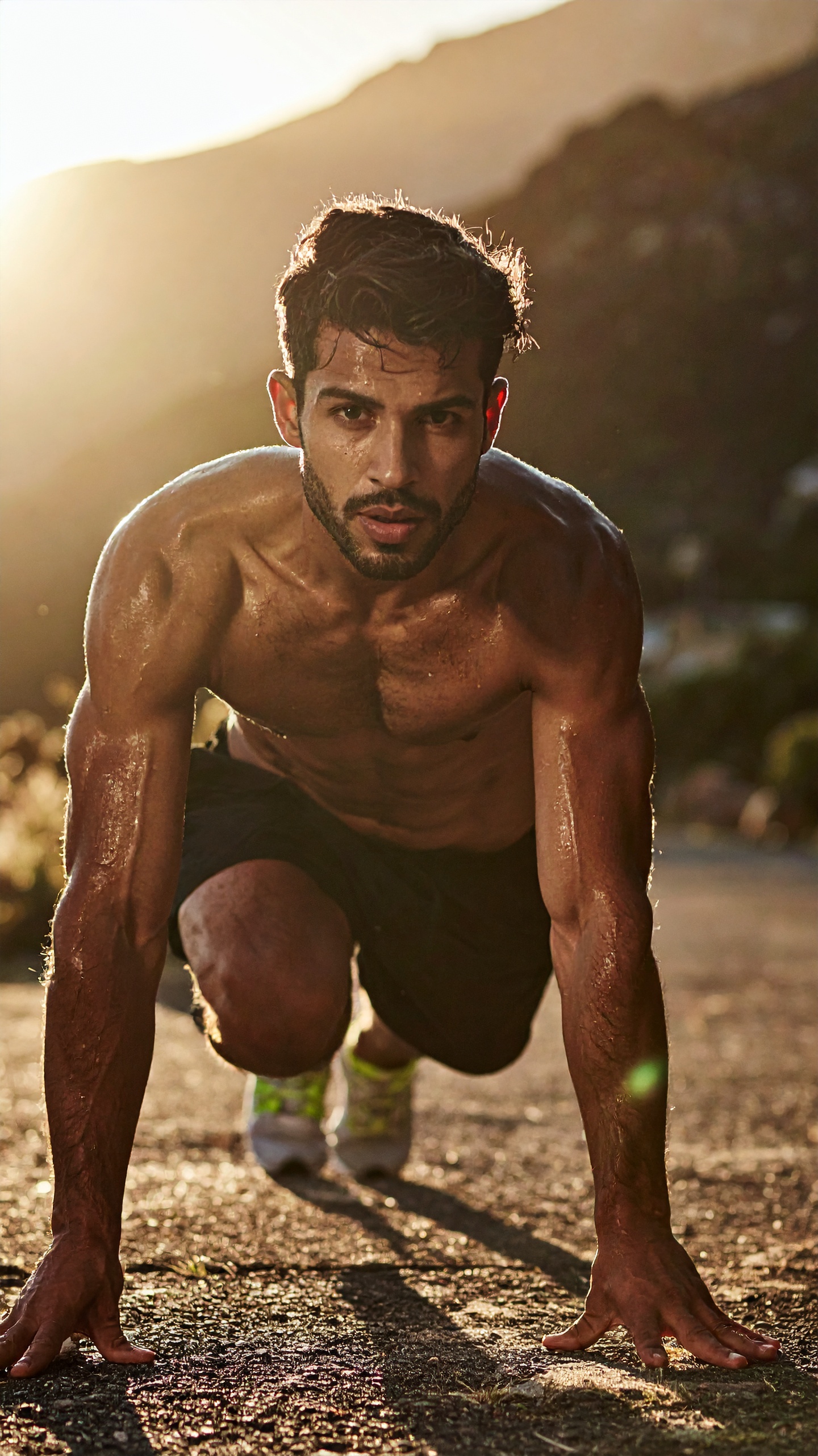 Atleta se prepara para corrida ao ar livre sob luz dourada do pôr do sol. Ideal para promover roupas esportivas, a imagem transmite energia e determinação. O suor no corpo e a expressão focada destacam o esforço físico e a força de vontade. Perfeito para campanhas de verão ou atividades ao ar livre, especialmente em locais montanhosos. A iluminação natural ressalta o dinamismo e a autenticidade da cena.