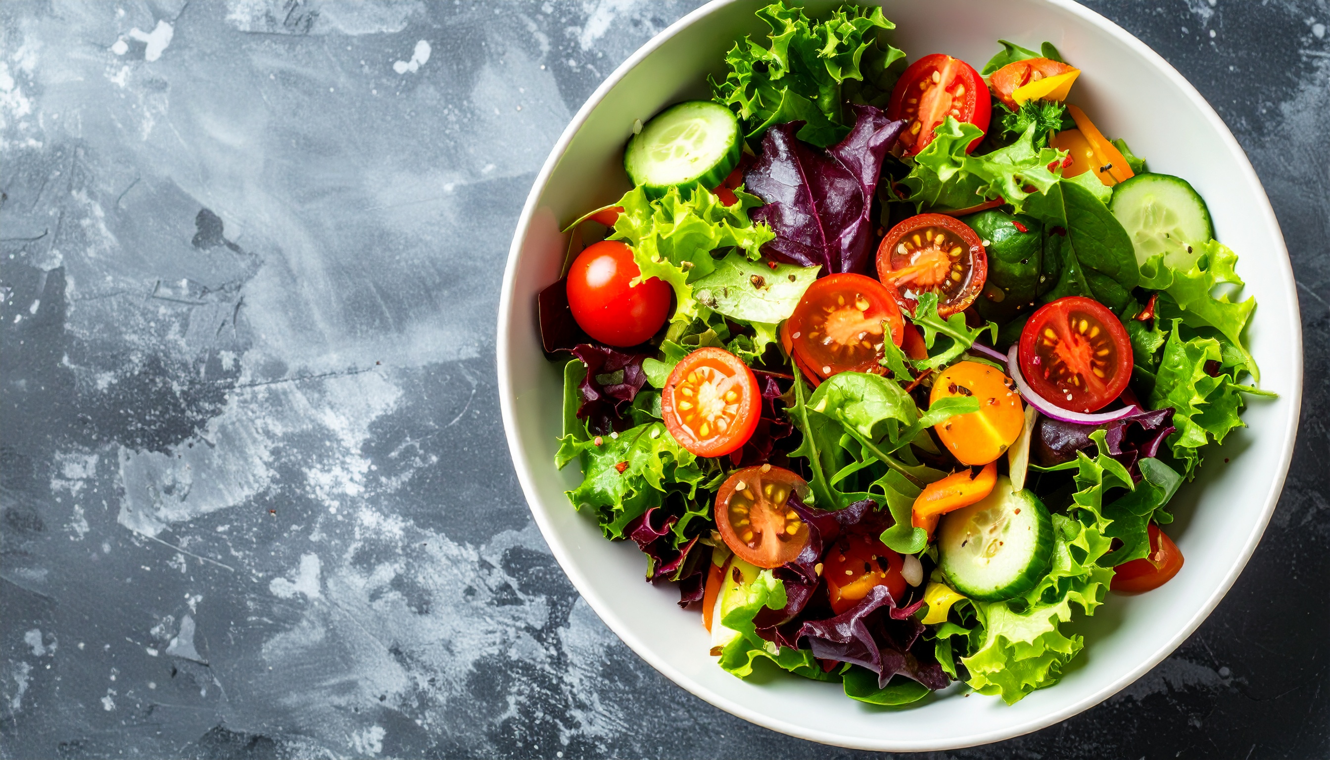 Salada colorida em tigela branca com folhas verdes, tomates cereja, pimentões e fatias de pepino sobre fundo cinza texturizado. Iluminação vibrante destaca frescor.