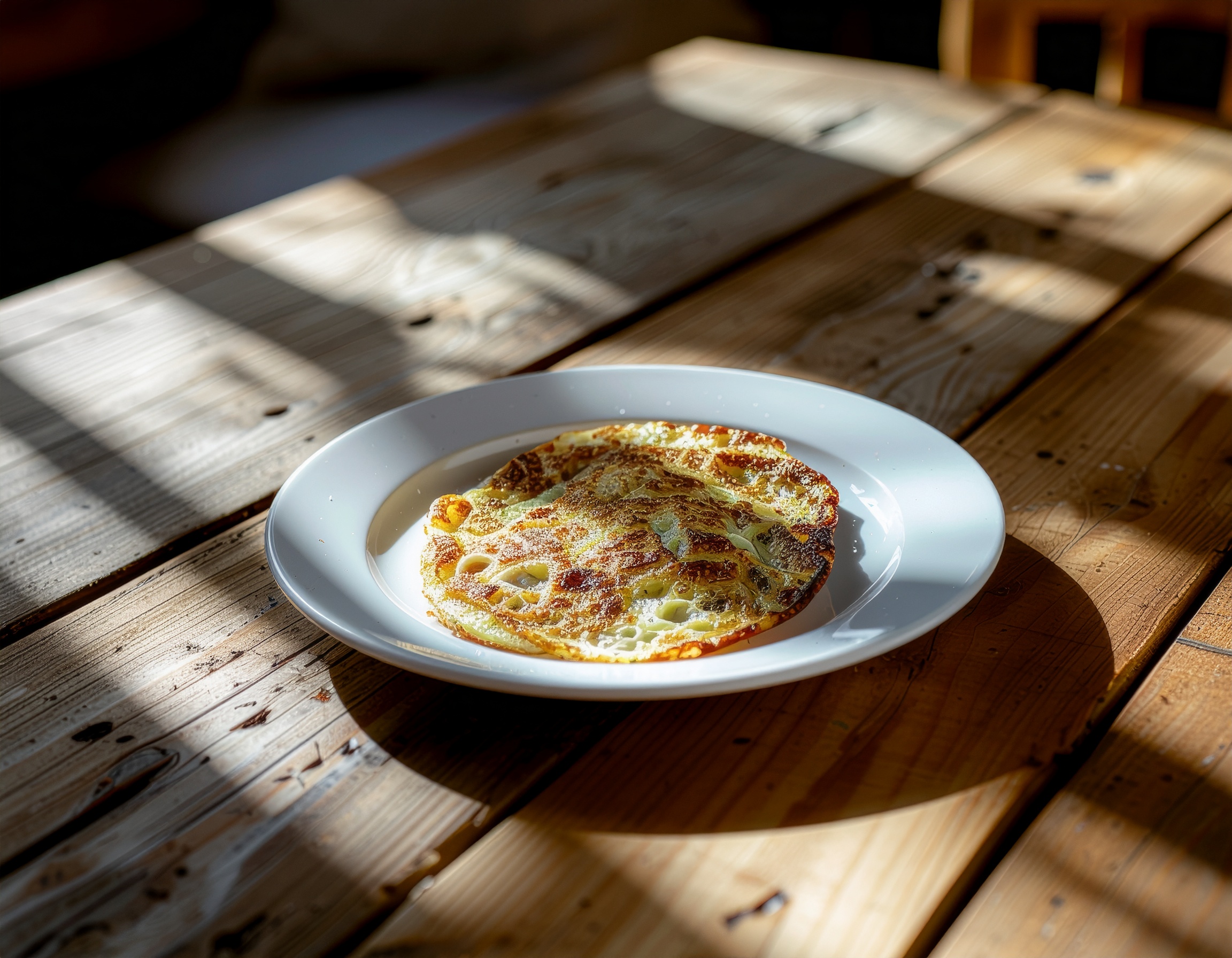 Panqueca dourada servida em um prato branco sobre uma mesa de madeira rústica. Ideal para um café da manhã ou brunch aconchegante, trazendo um toque caseiro às refeições. A textura crocante e a apresentação simples destacam-se sob a luz suave do dia. Perfeita para ser saboreada em ambientes descontraídos, como uma cozinha ou café acolhedor. A combinação de elementos naturais e culinários oferece um apelo visual encantador.