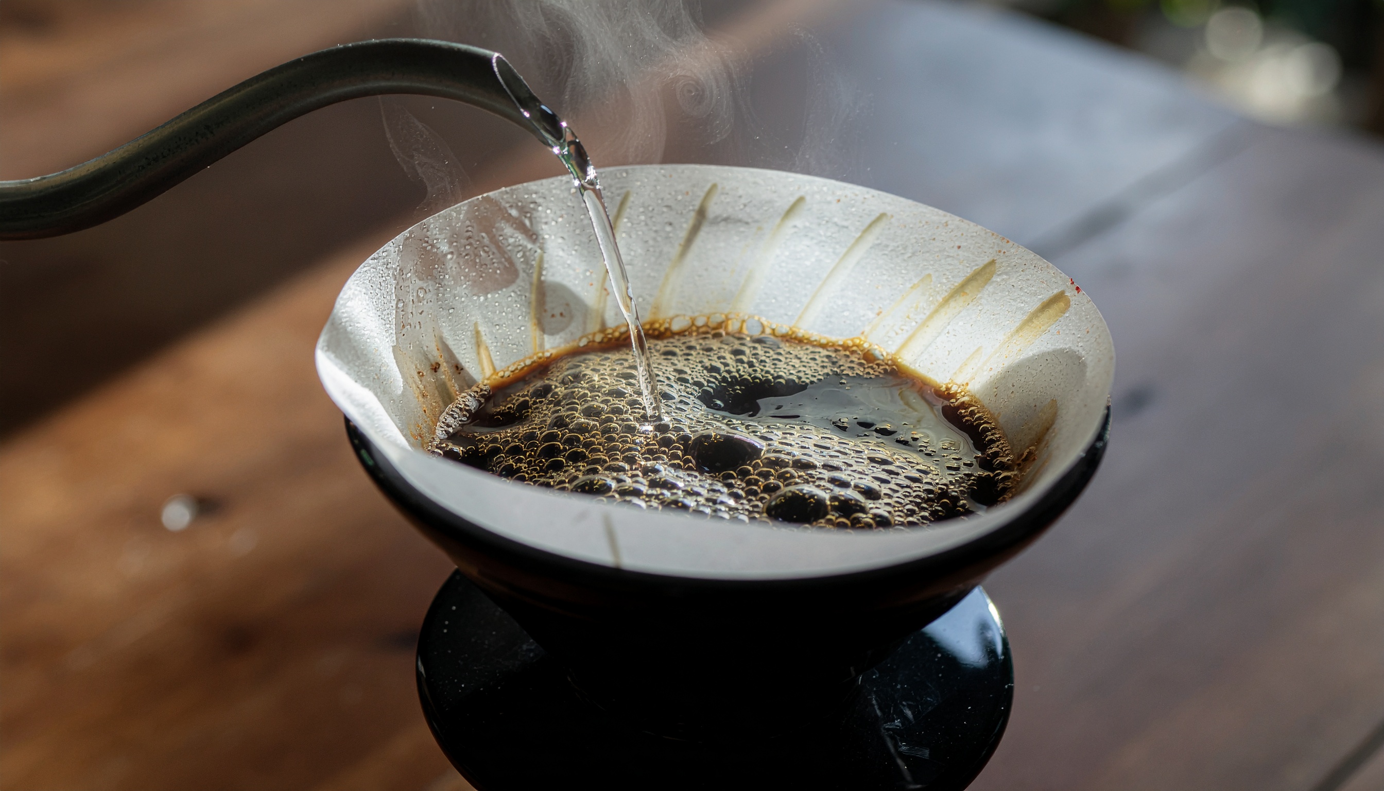 Preparação de café por método de filtragem manual, com jarro despejando água quente sobre grãos moídos em filtro de papel, criando espuma cremosa. Luz suave realça o contraste entre o líquido escuro e o filtro, capturando vapor sutil. Composição dinâmica com foco no filtro e fundo desfocado, proporcionando uma sensação de ritual matinal tranquilo.