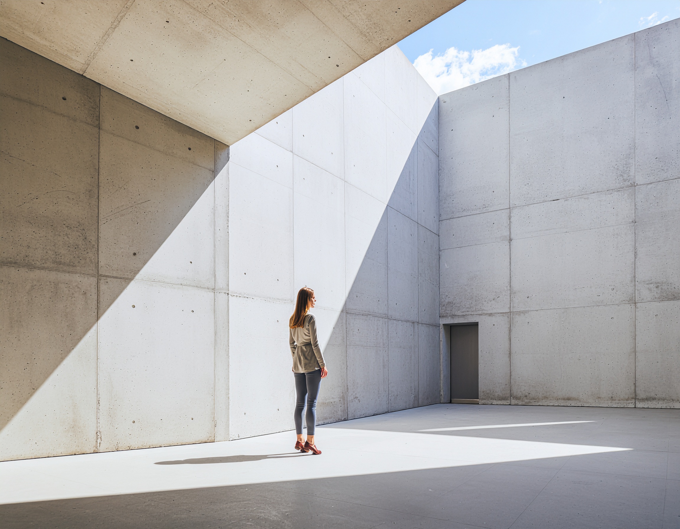 A woman stands in a modern concrete courtyard with sharp sunlight casting geometric shadows