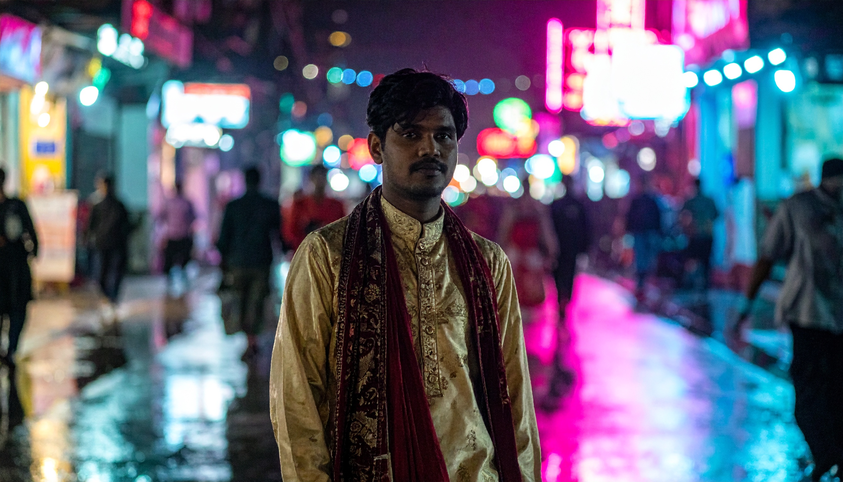 A man in traditional attire stands on a vibrant, neon-lit street at night