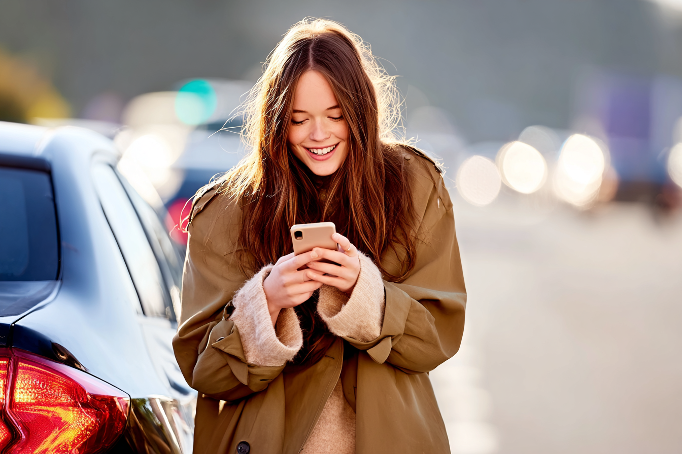 Mulher sorridente usando casaco bege segura smartphone em uma rua iluminada. O fundo desfocado sugere movimento urbano e luzes de tráfego. Iluminação suave realça o cabelo, criando uma atmosfera alegre e espontânea. A composição centraliza a figura, com ênfase em sua expressão de felicidade. O ângulo frontal e foco nítido destacam detalhes da roupa e do dispositivo.