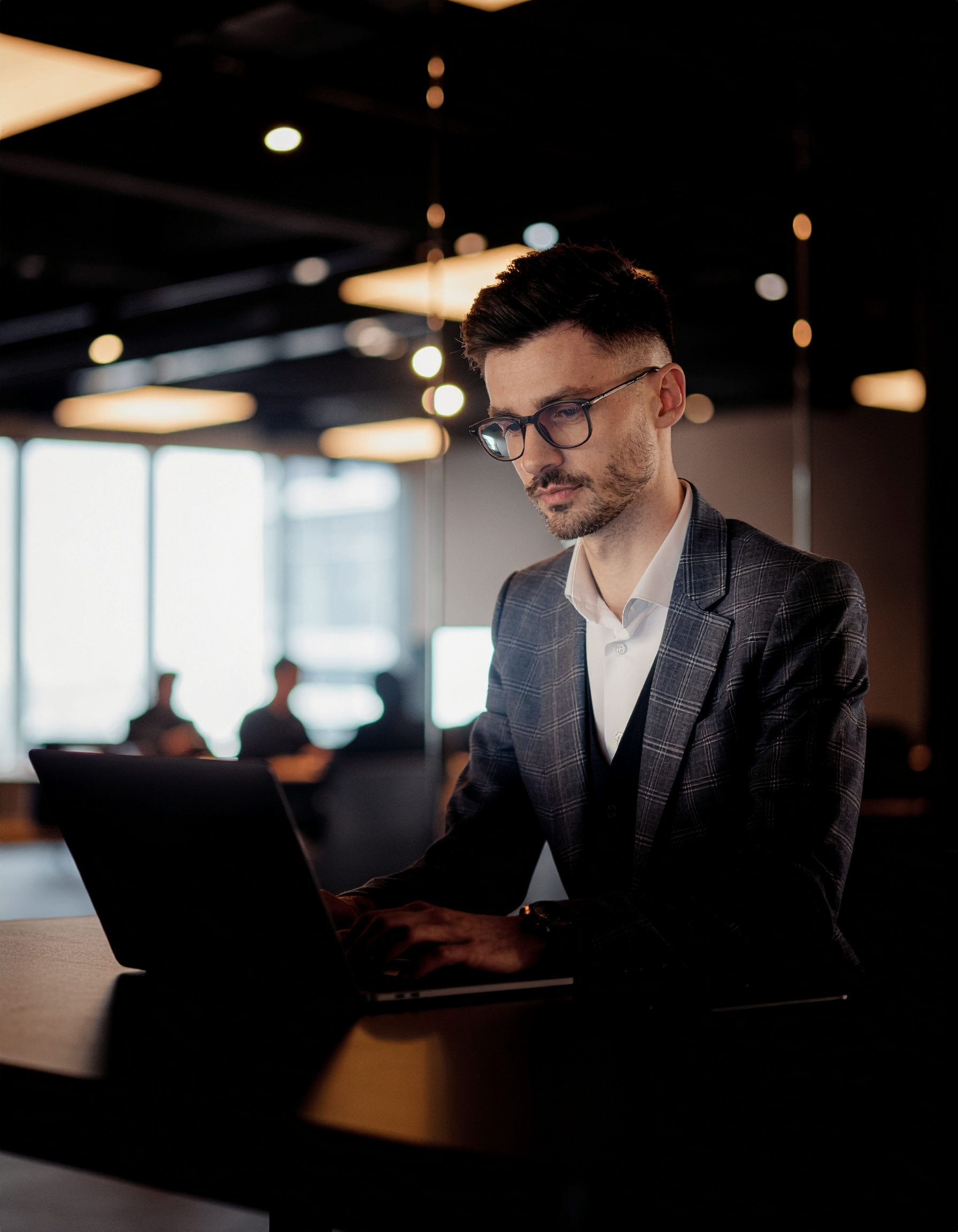 Homem sentado em um ambiente de escritório moderno, trabalhando em um laptop. A iluminação suave e difusa confere um tom profissional e focado. O fundo desfocado com silhuetas de pessoas sugere um espaço de coworking. O homem veste um terno xadrez e óculos, concentrado em sua tarefa. A composição é elegante, com uso de luz natural e ângulos retos, destacando a seriedade do ambiente de trabalho.